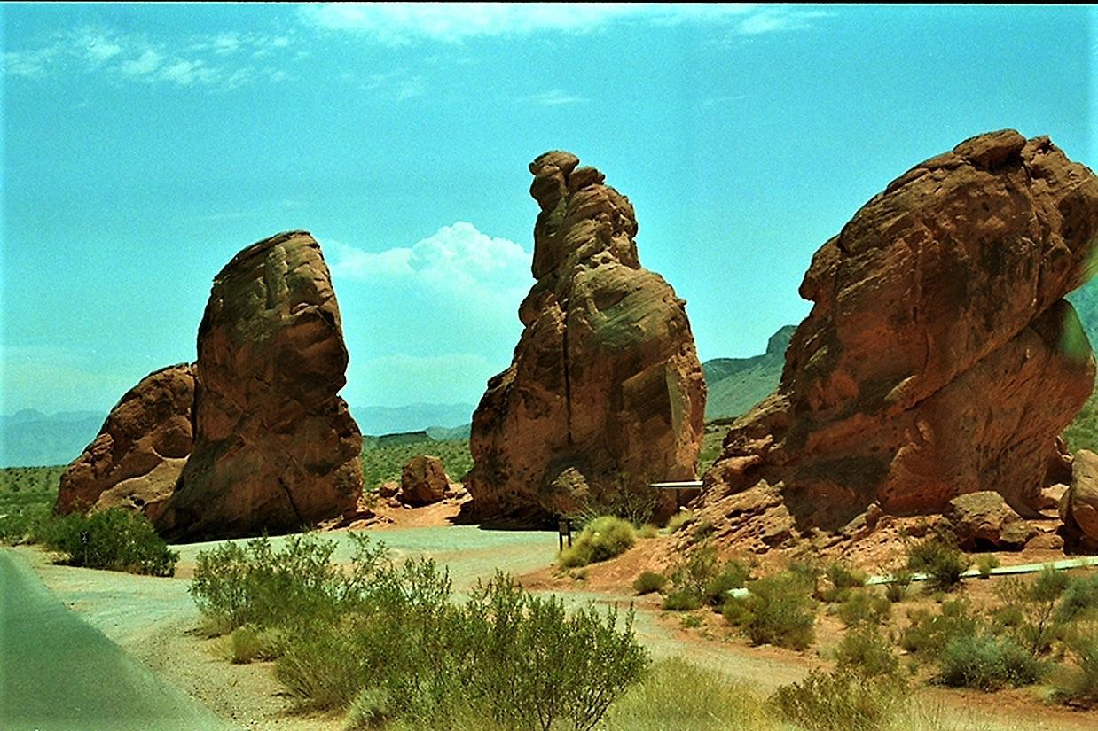 Three large red rock formations beside a road, under a bright blue sky.