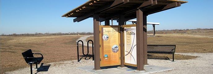 Covered trail information kiosk, bench, and bike rack in a sunny, open field.