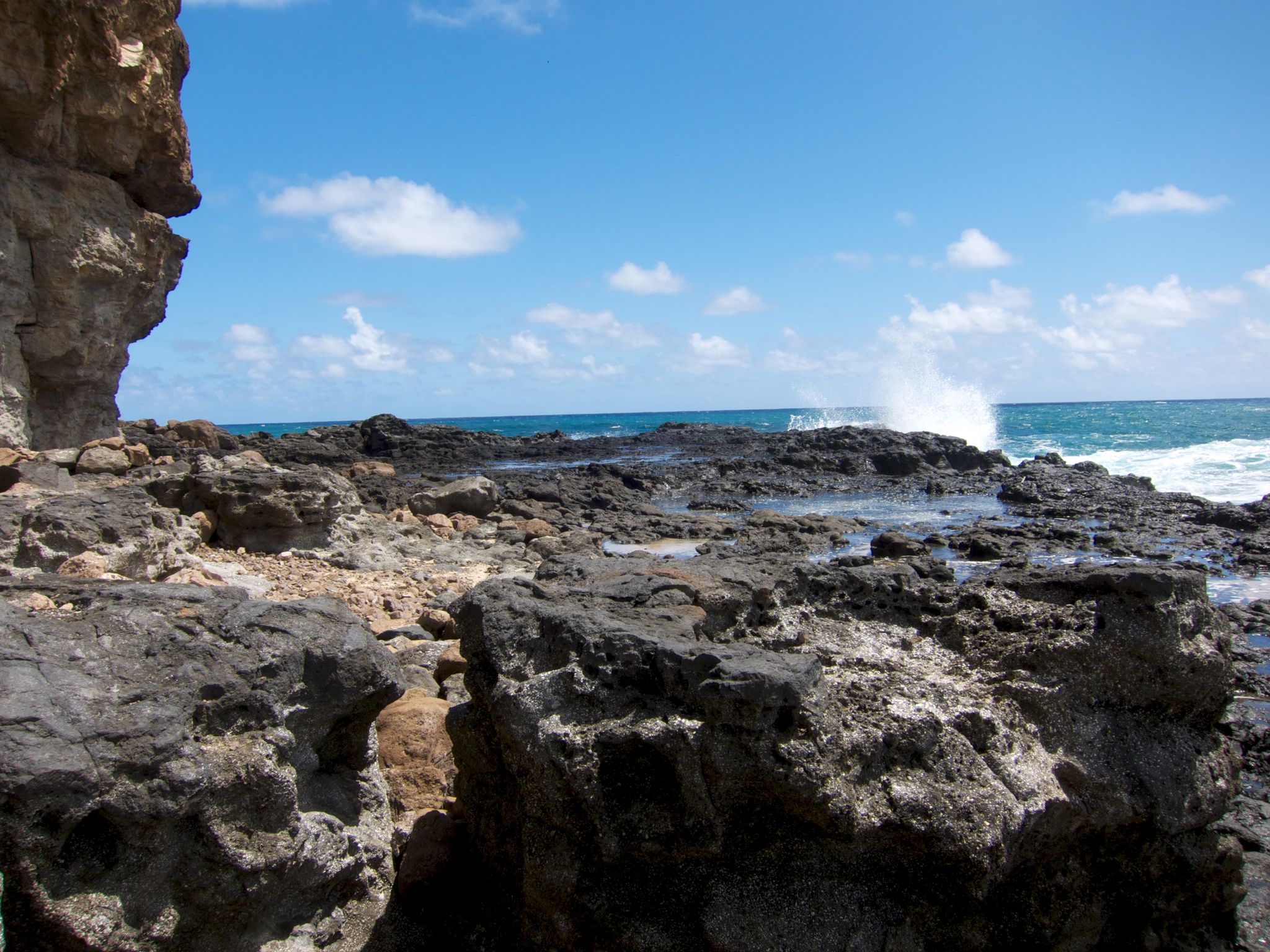 Waves crash against dark, jagged rocks under a bright blue sky. - Mokolea Point Trail