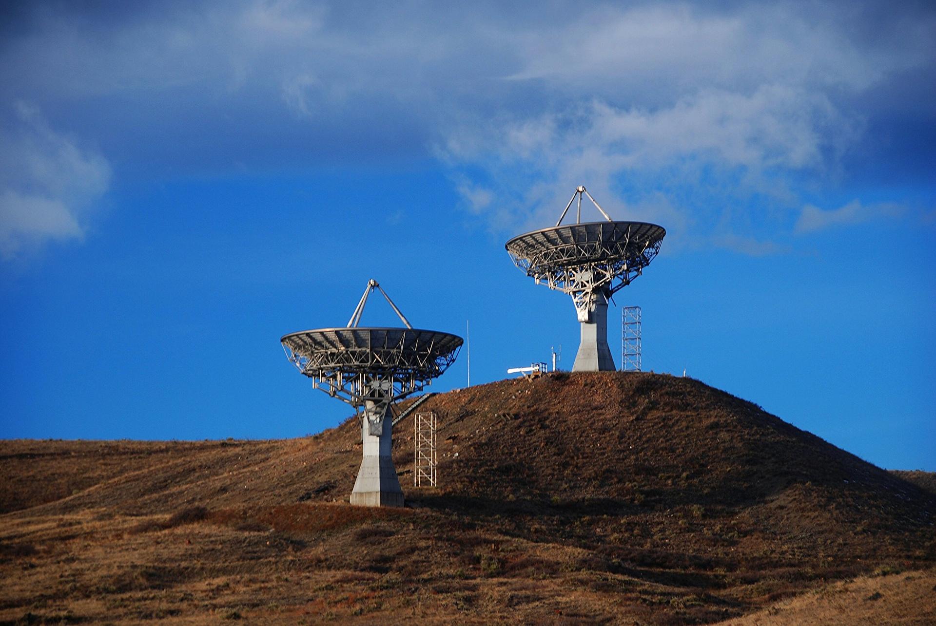 Two large satellite dishes on a barren hill under a dramatic blue sky.
