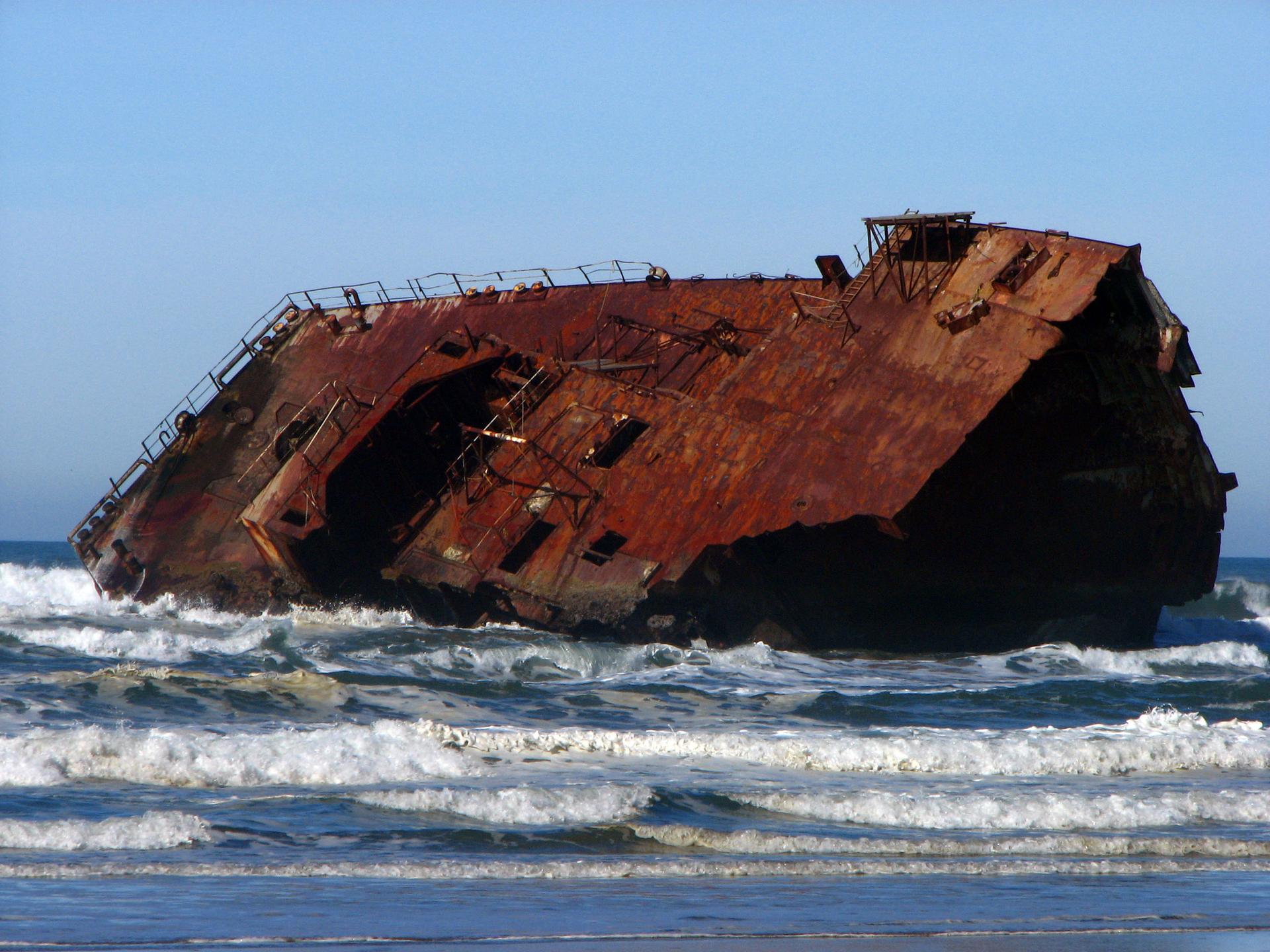 A large, rusty shipwreck sits partially submerged in ocean waves on a bright day.