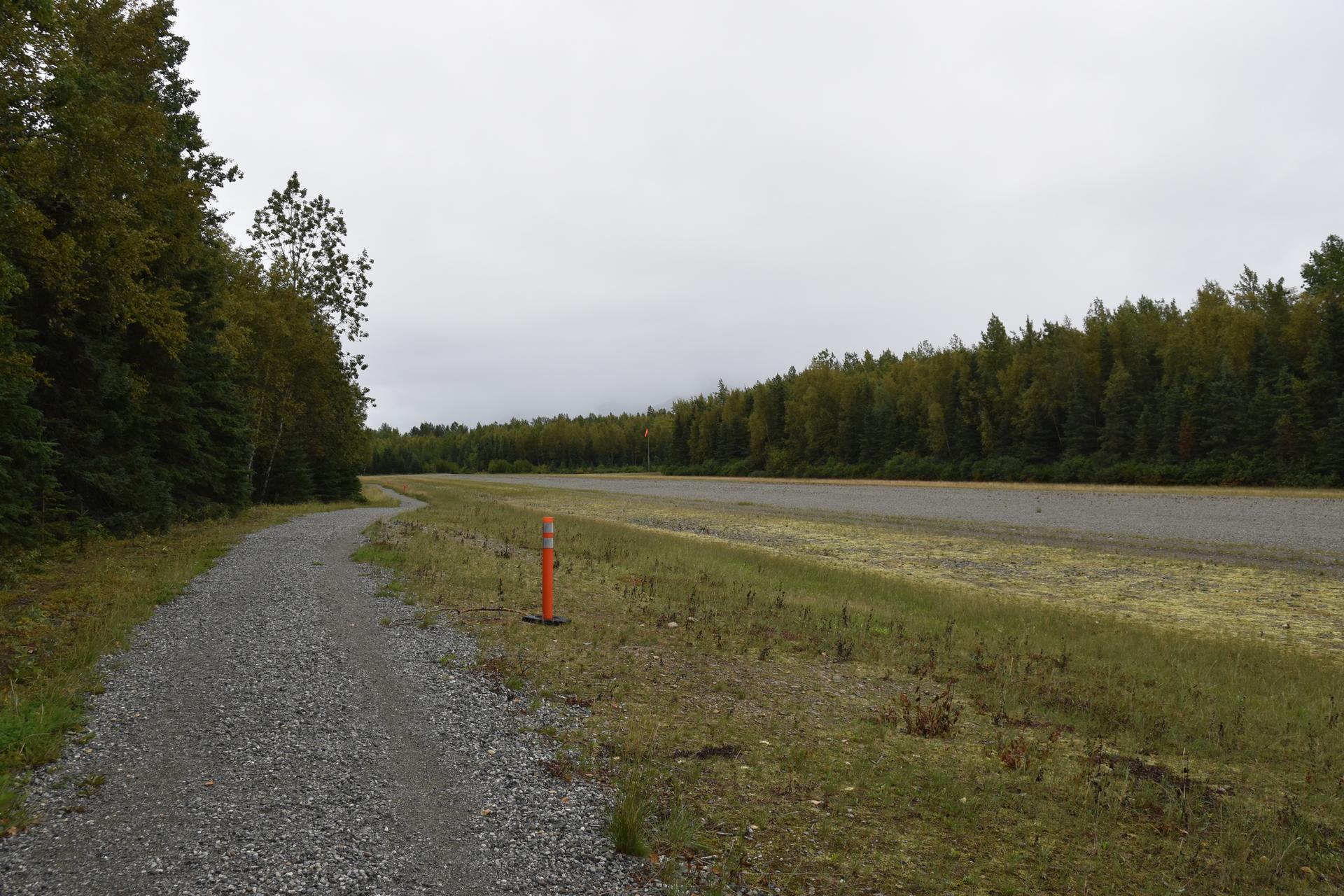 Gravel path beside a wide grassy field, bordered by dense forest under a cloudy sky.
