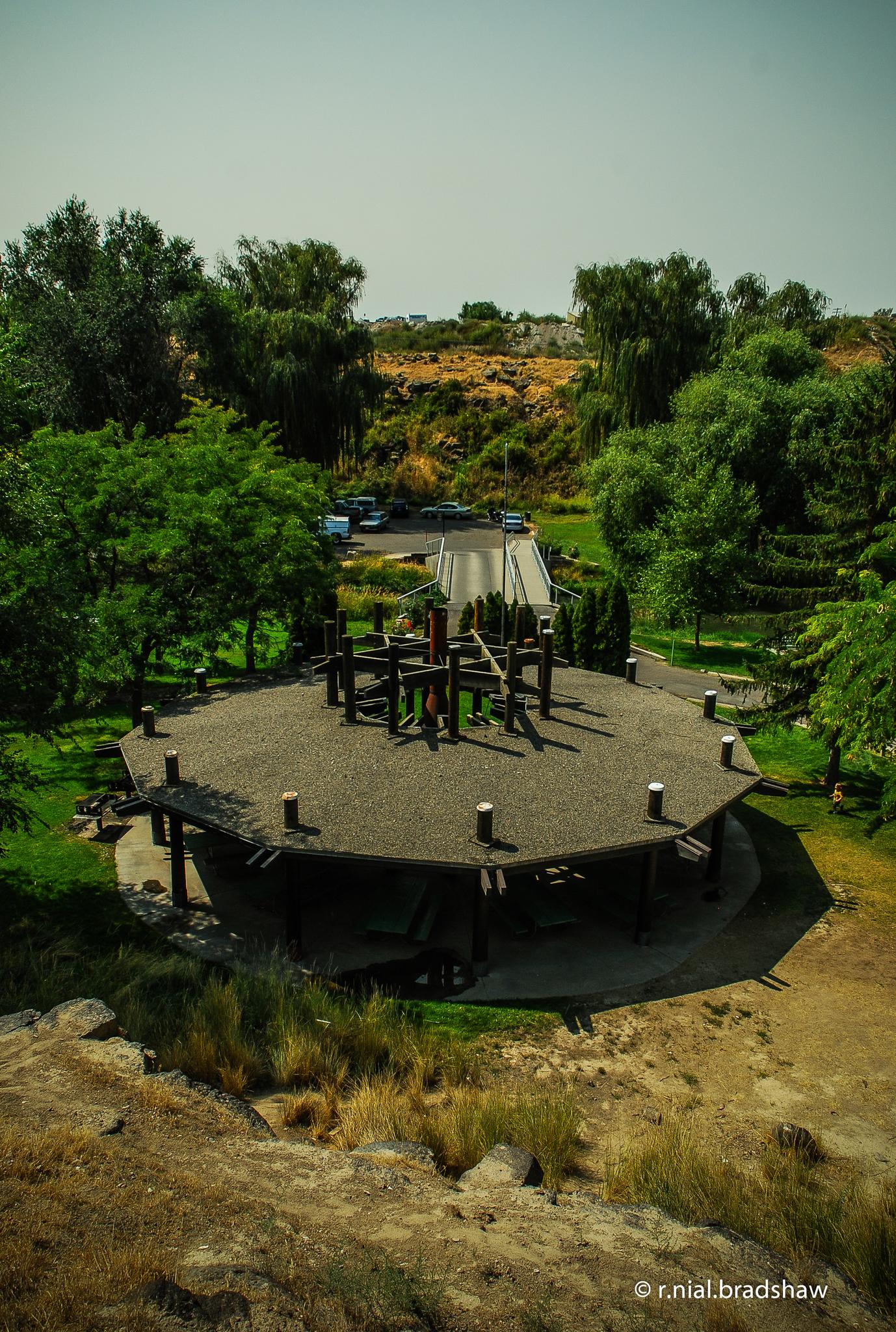 Overhead view of a large, dark-roofed pavilion amidst green trees and a road.