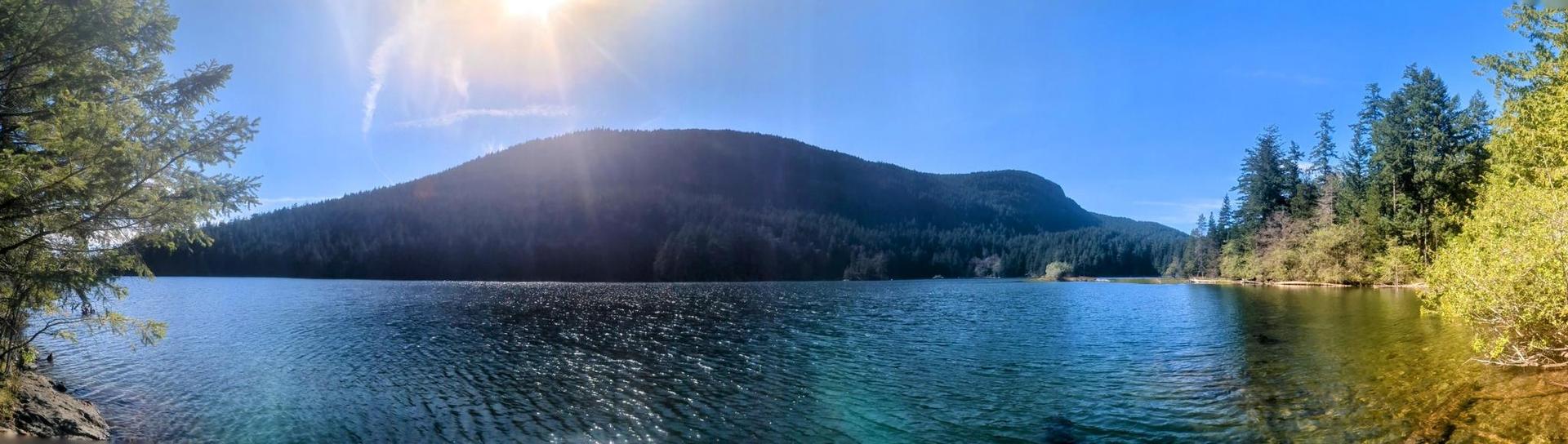 Panoramic view of a clear lake, dense forest, and mountain under bright sun.