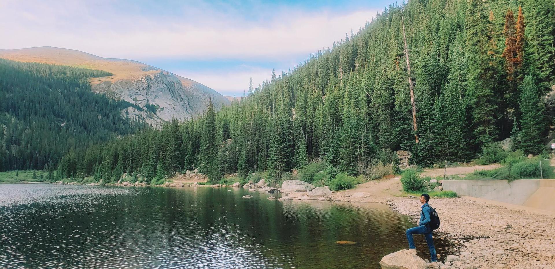 Person with backpack stands on rocks by a lake, surrounded by tall pine trees.