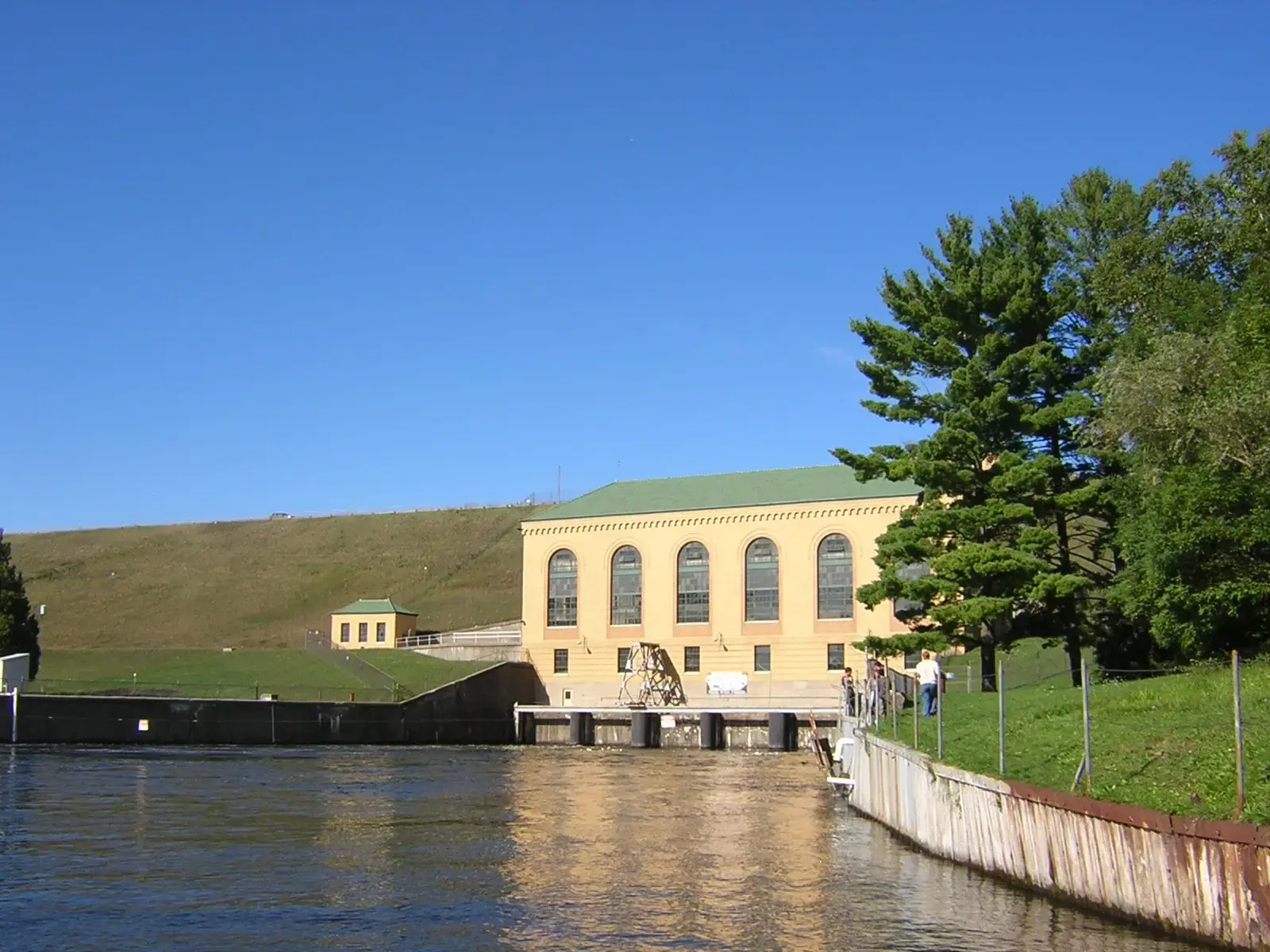Yellow building by water with a grassy hill under a clear blue sky.