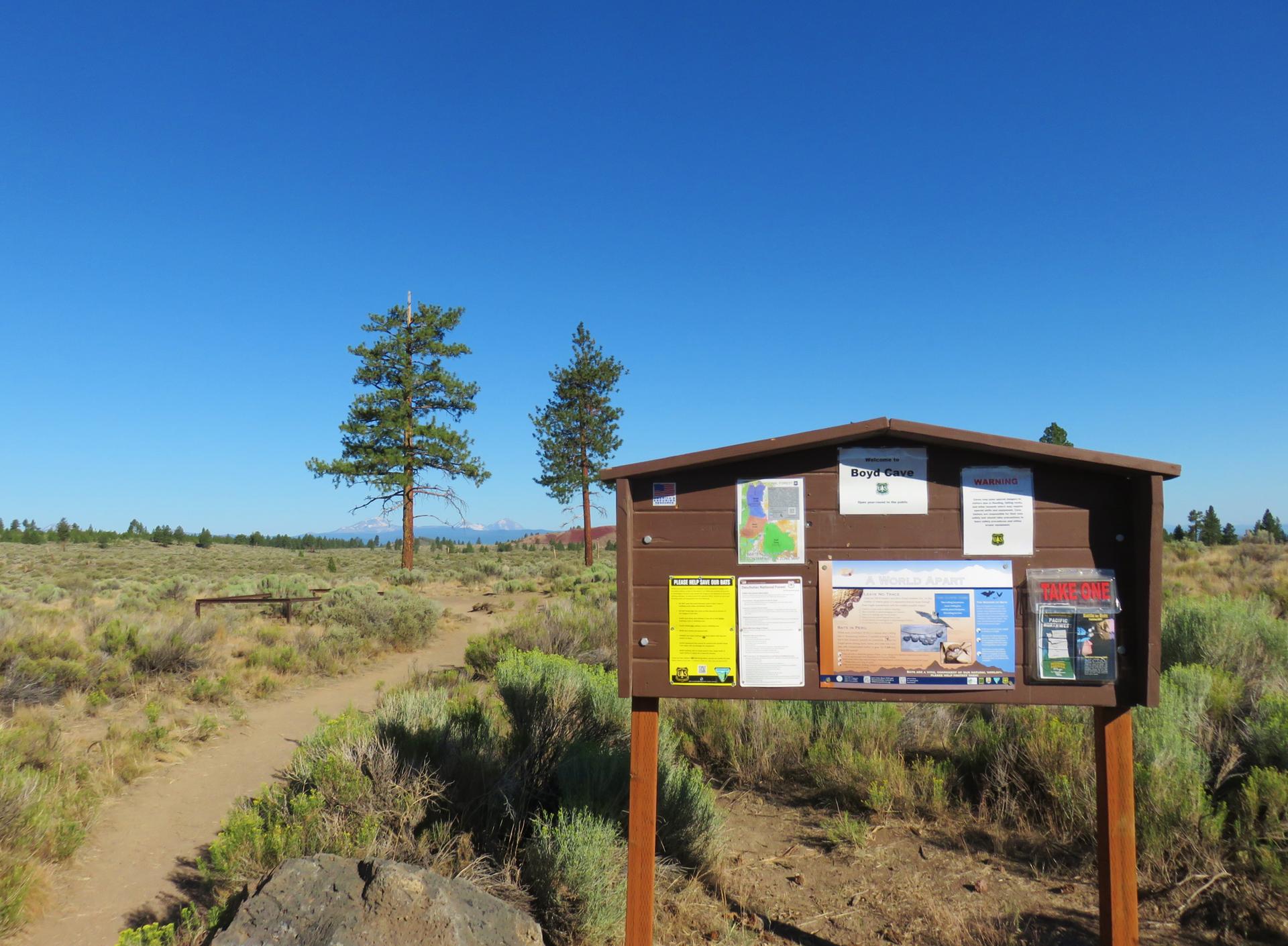 Wooden information board beside a dirt path in a sunny, arid landscape.