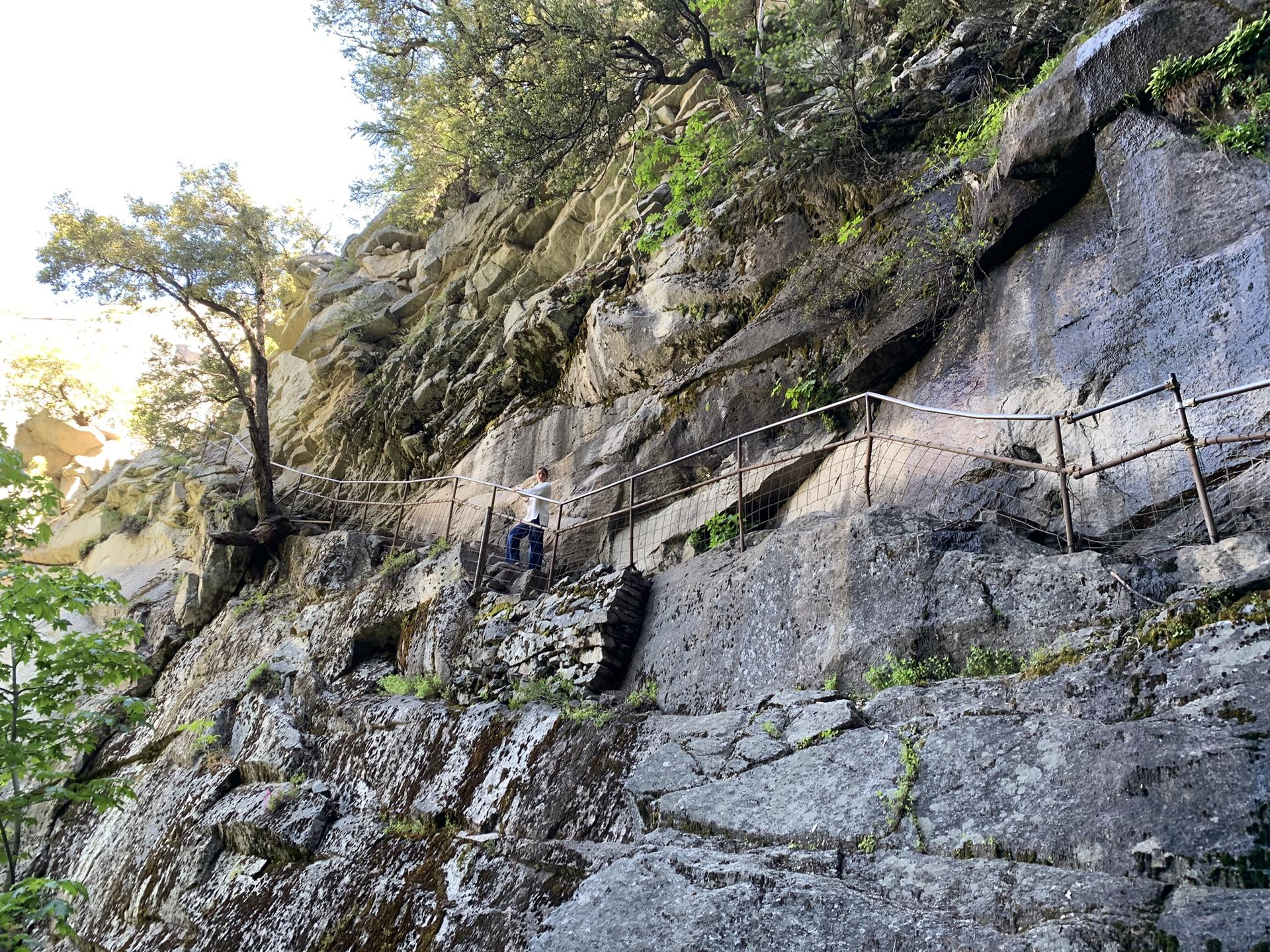 A person walks on a narrow, rocky path with a railing along a cliff.