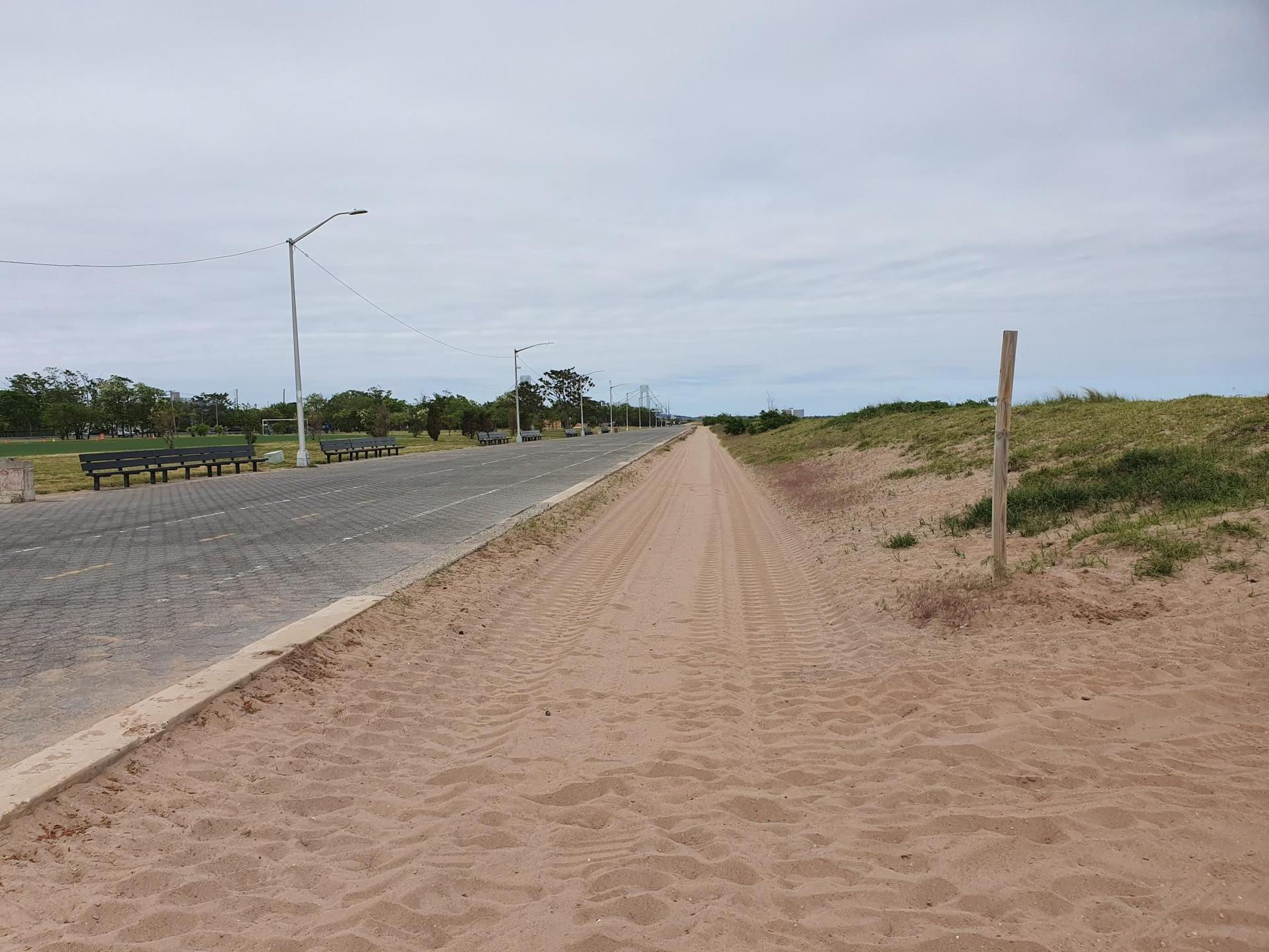 Sandy path next to a paved road with benches, under a cloudy sky.