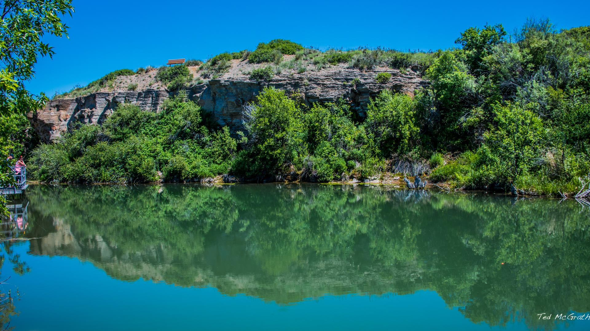 Bright sunny day with calm water reflecting green trees and rocky cliffs.