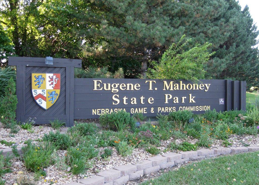 Large wooden sign with a crest, surrounded by green trees and shrubs.