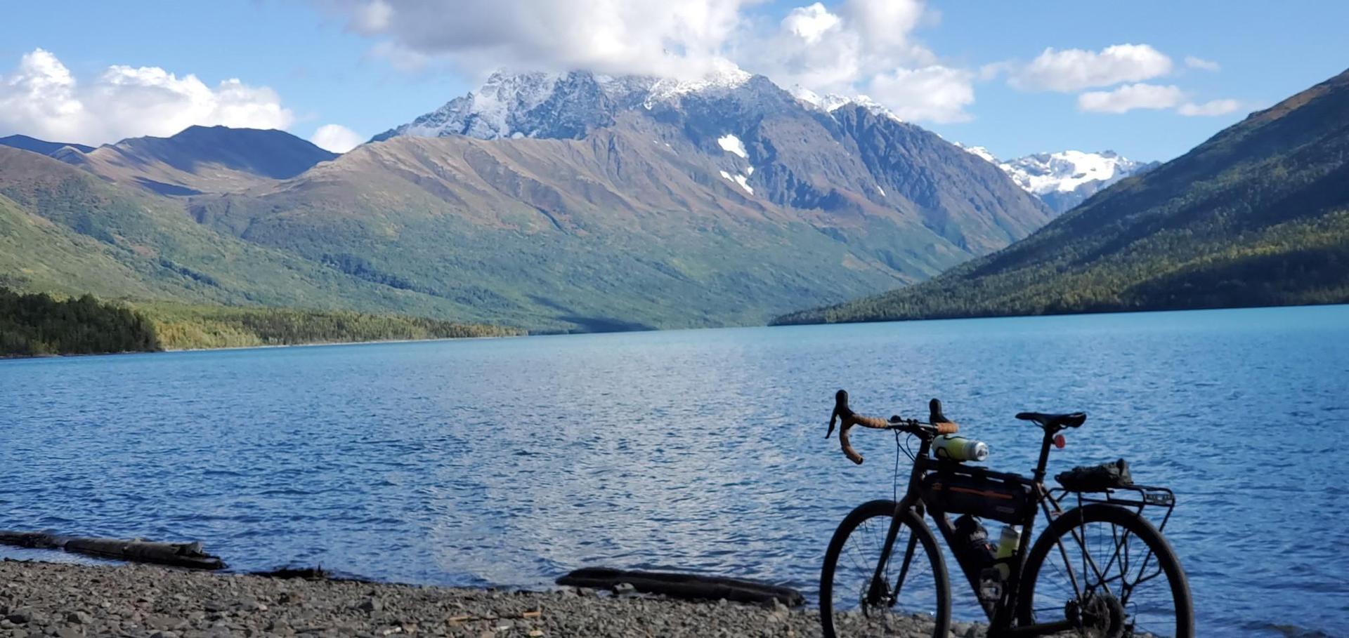 A bicycle rests on a rocky shore beside a turquoise lake and snow-capped mountains.