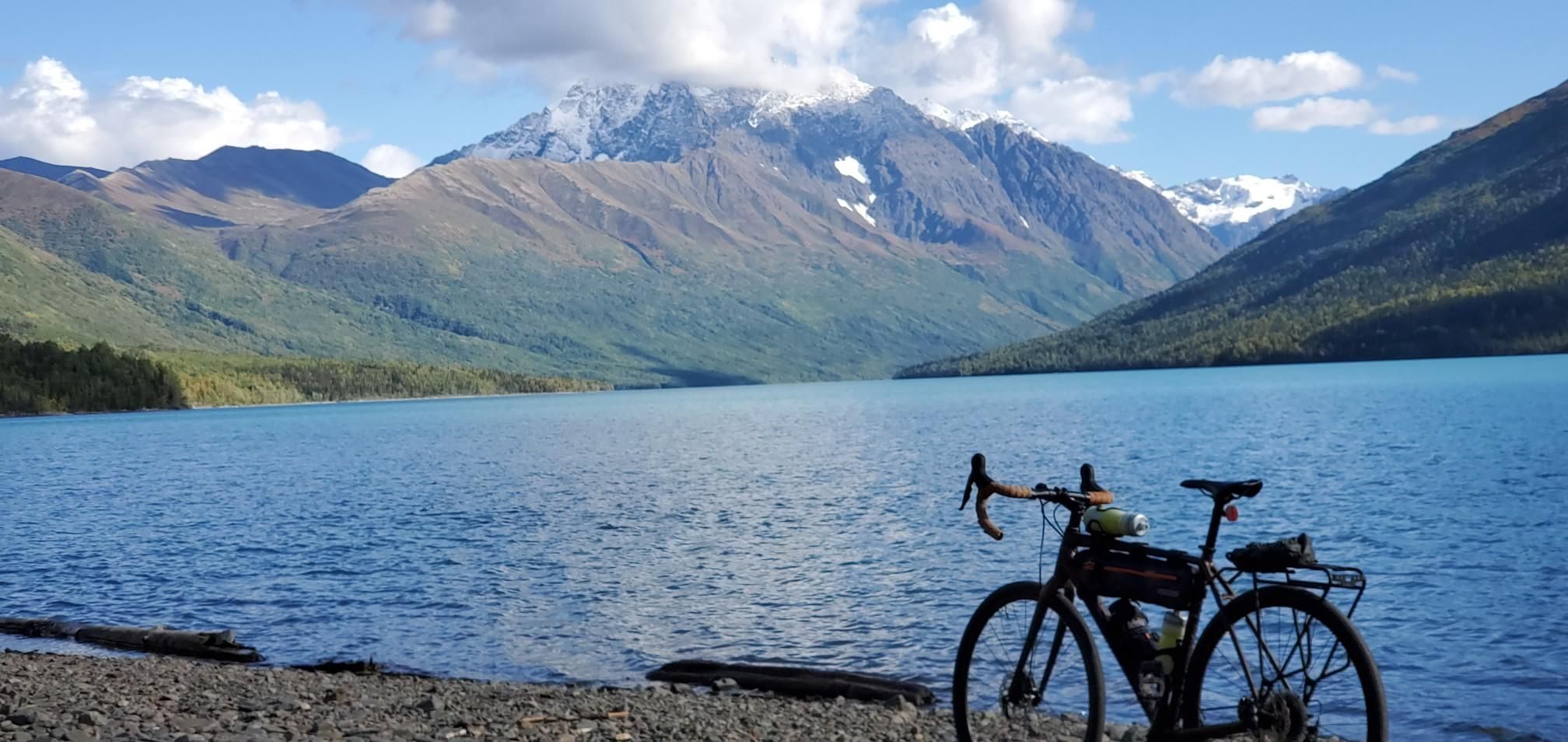 A bicycle rests on a rocky shore beside a turquoise lake and snow-capped mountains. - Twin Peaks Trail