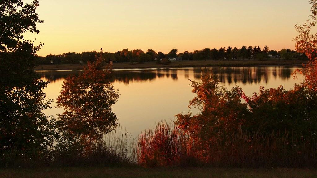 Warm sunset glow over a calm lake, framed by autumn-colored trees.