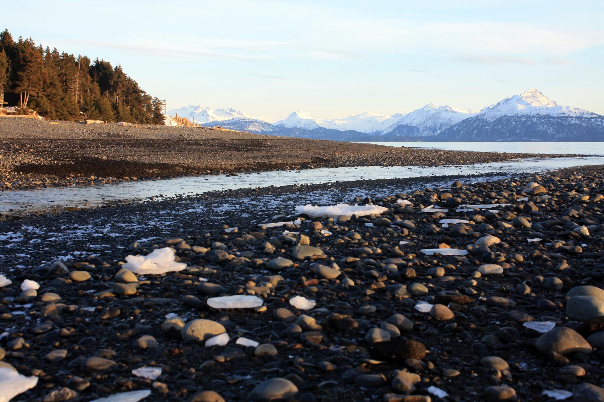 Rocky beach with ice chunks, a stream, and distant snowy mountains at sunset.