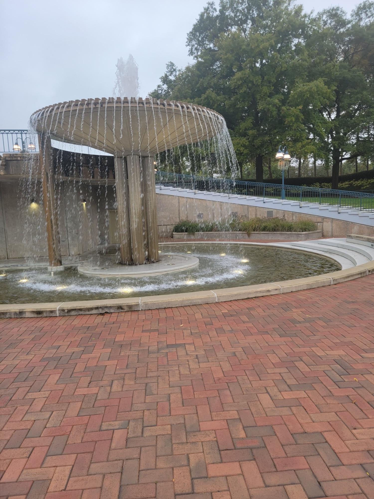 Large, tiered fountain with cascading water on a brick plaza under an overcast sky.
