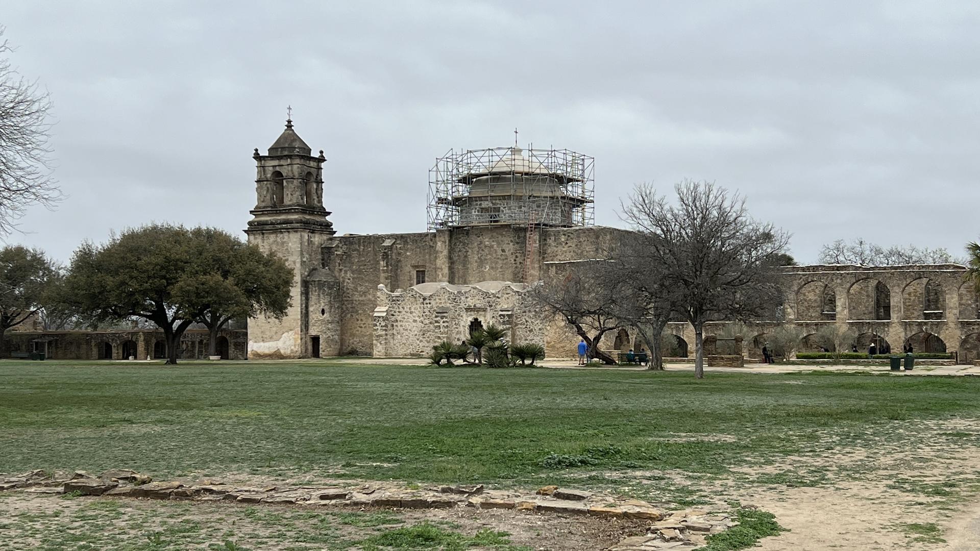 Large stone building with a tower and scaffolding, green lawn, cloudy day.
