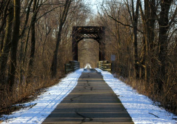 Sunny winter path with snow leading to a dark, arching bridge.