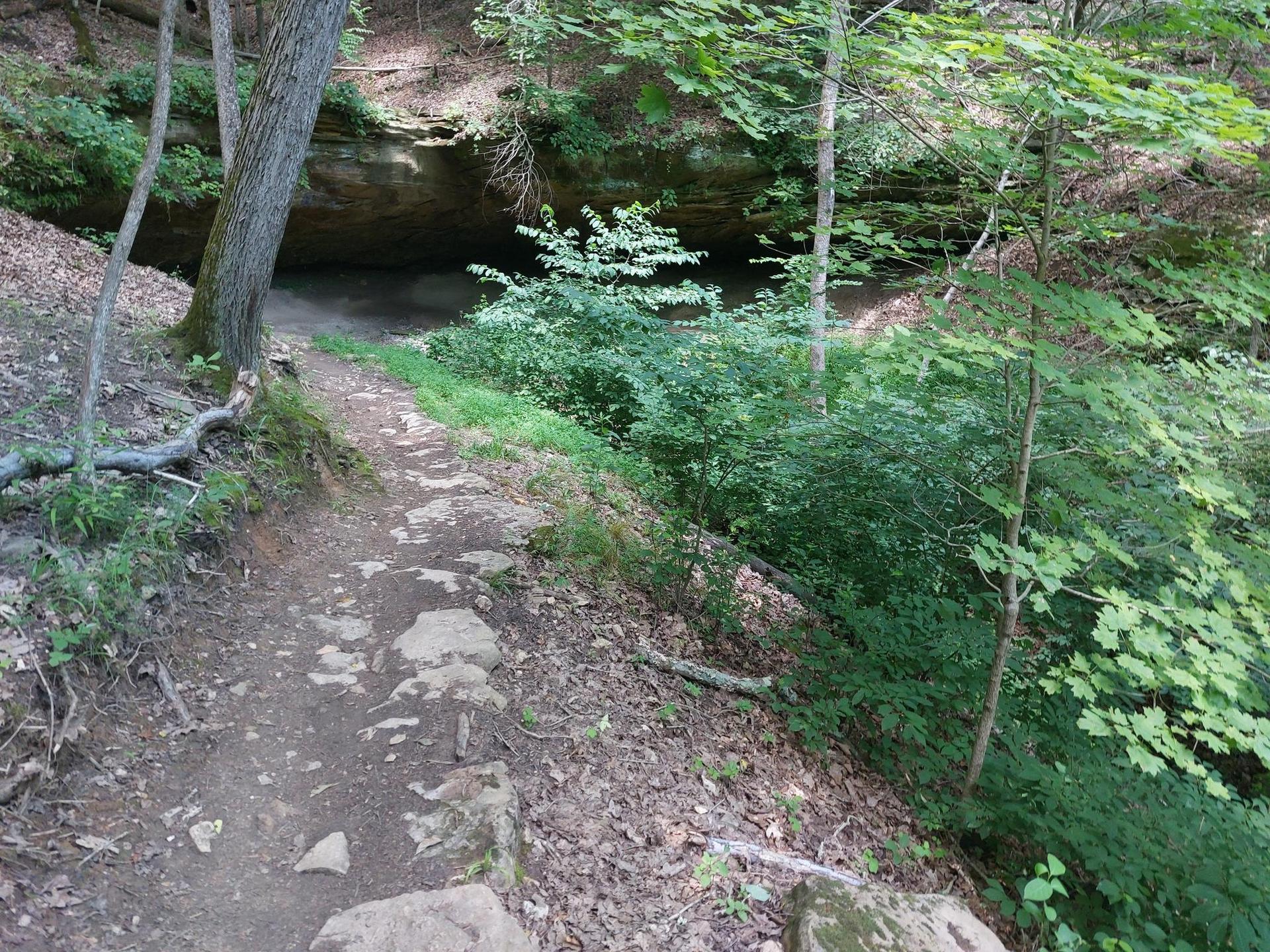 A rocky dirt path leads into a shaded rock overhang surrounded by green foliage.