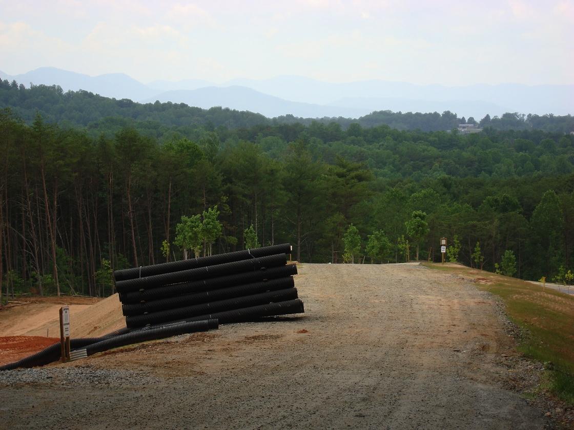Stack of black pipes on a dirt road with forested mountains under an overcast sky.