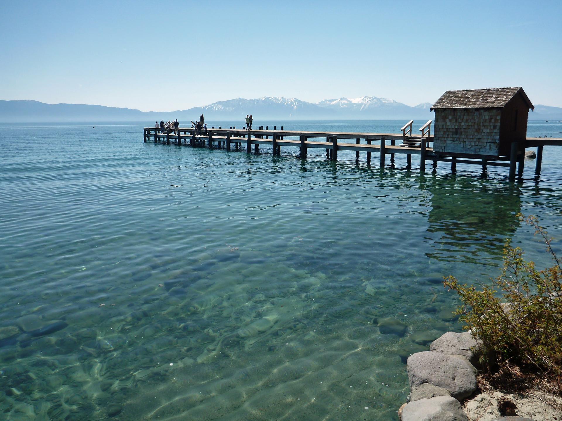 Long wooden pier with a small building over clear blue water and distant mountains.