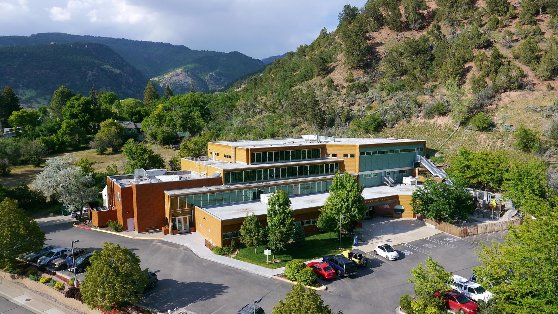 Modern building with parking lot, surrounded by green trees and distant mountains.