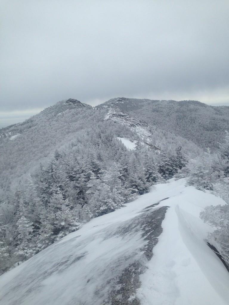 Snow and frost cover a mountain ridge with distant peaks under gray sky.