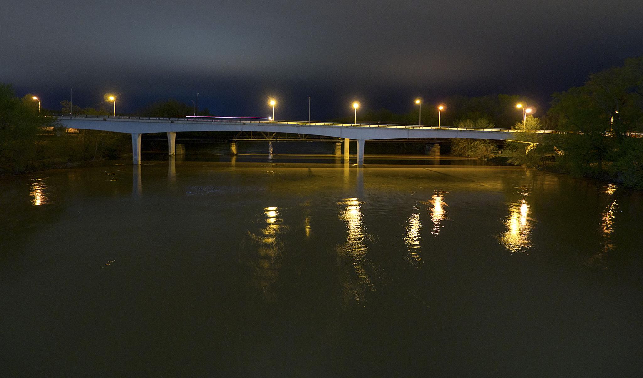 Brightly lit bridge spanning dark water at night, with reflections.