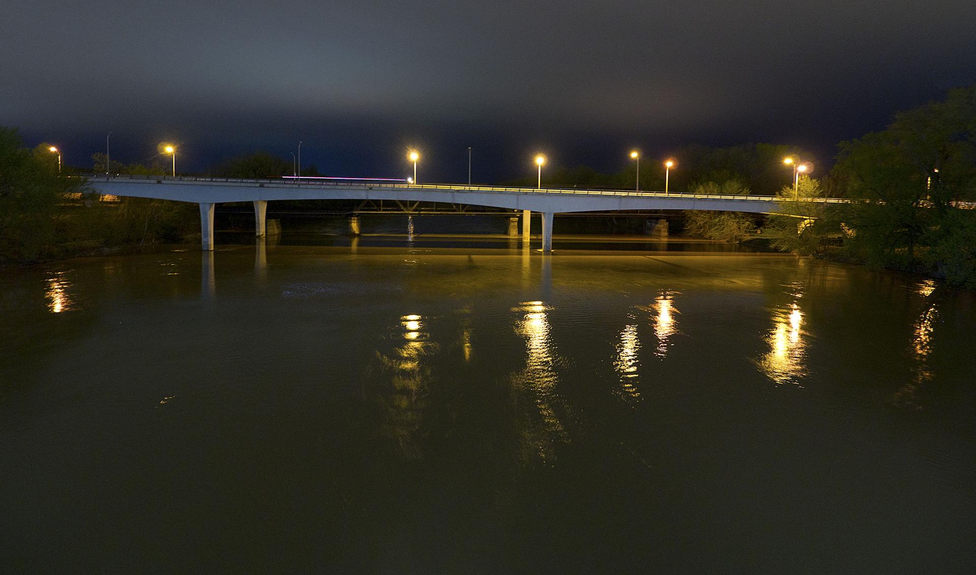 Brightly lit bridge spanning dark water at night, with reflections.