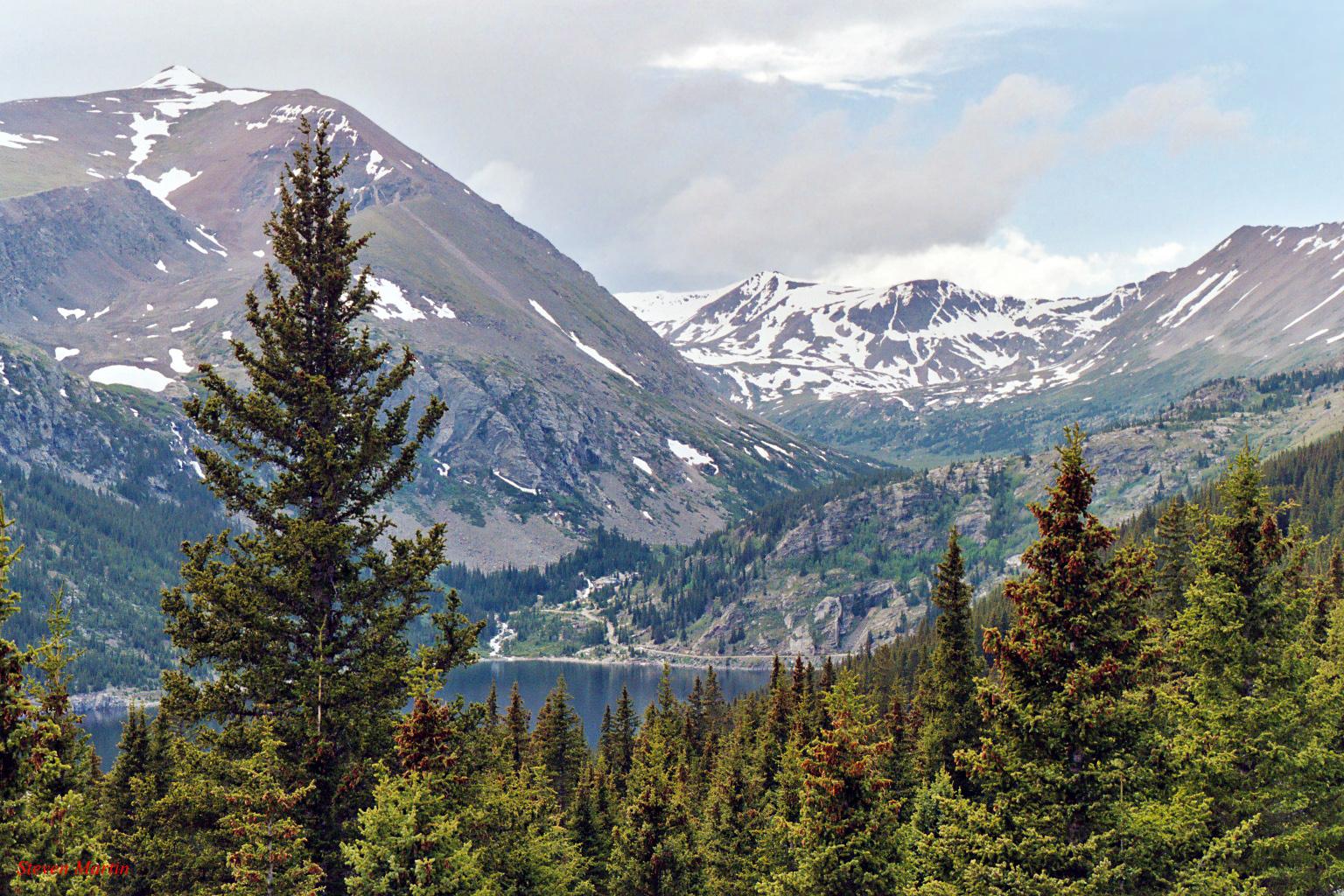 Pine trees frame a calm lake with snow-capped mountains under an overcast sky.