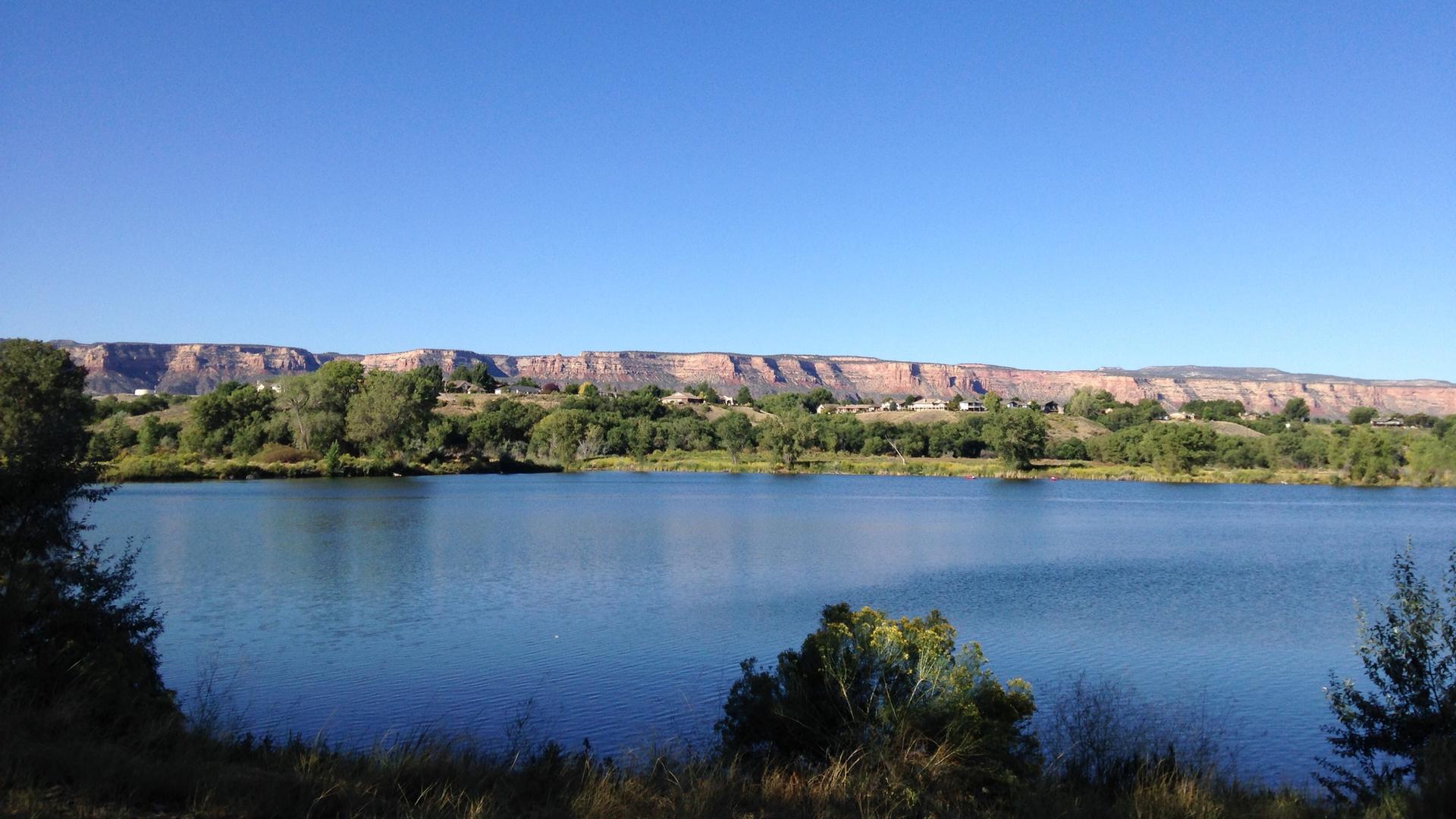 Calm lake reflecting clear blue sky, with green trees and distant cliffs.