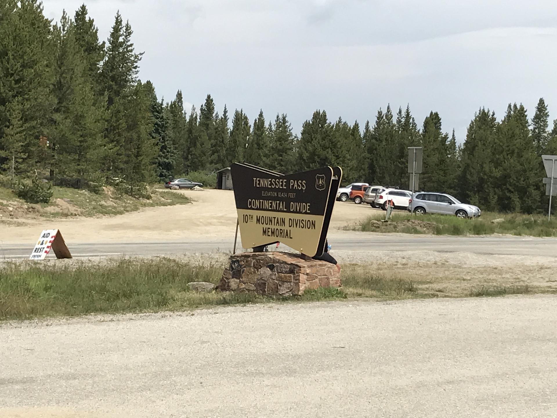 A brown roadside sign on a gravel area with parked cars and pine trees.