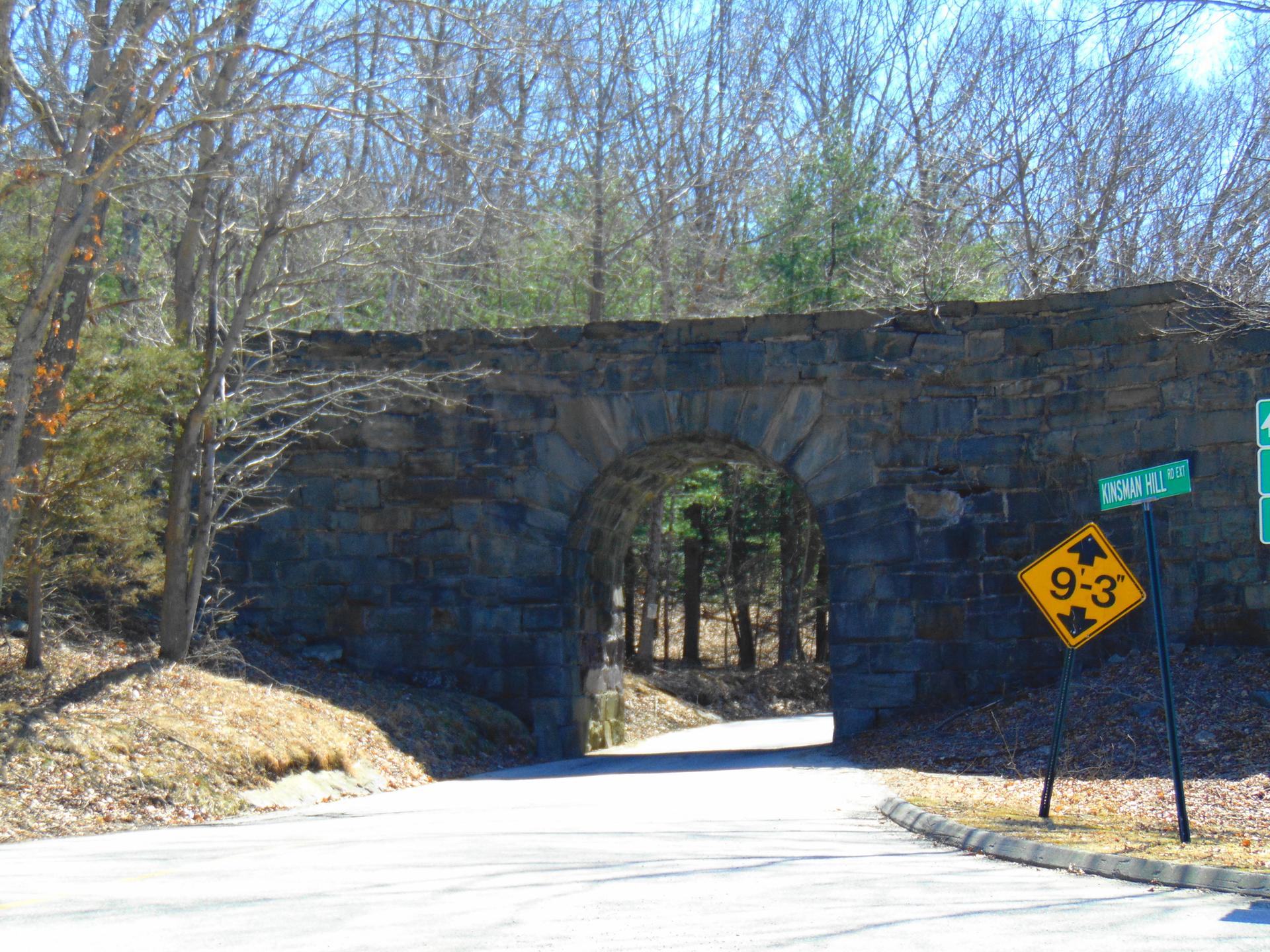 Stone arch bridge over a road, surrounded by bare trees on a sunny day.