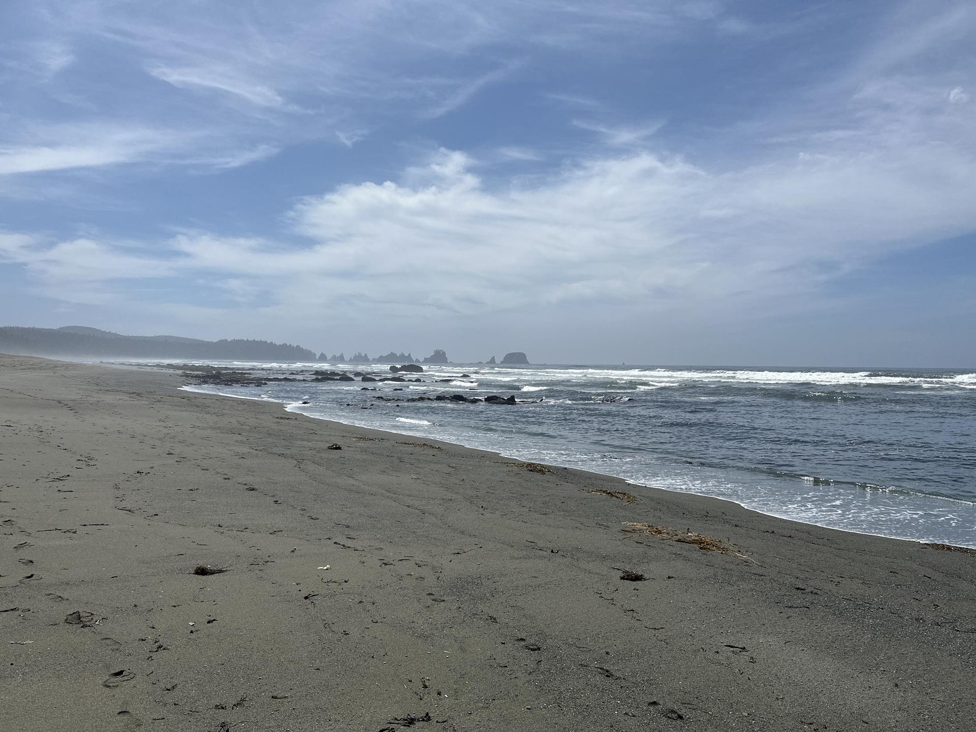 Wide sandy beach with ocean waves and distant sea stacks under a cloudy sky.