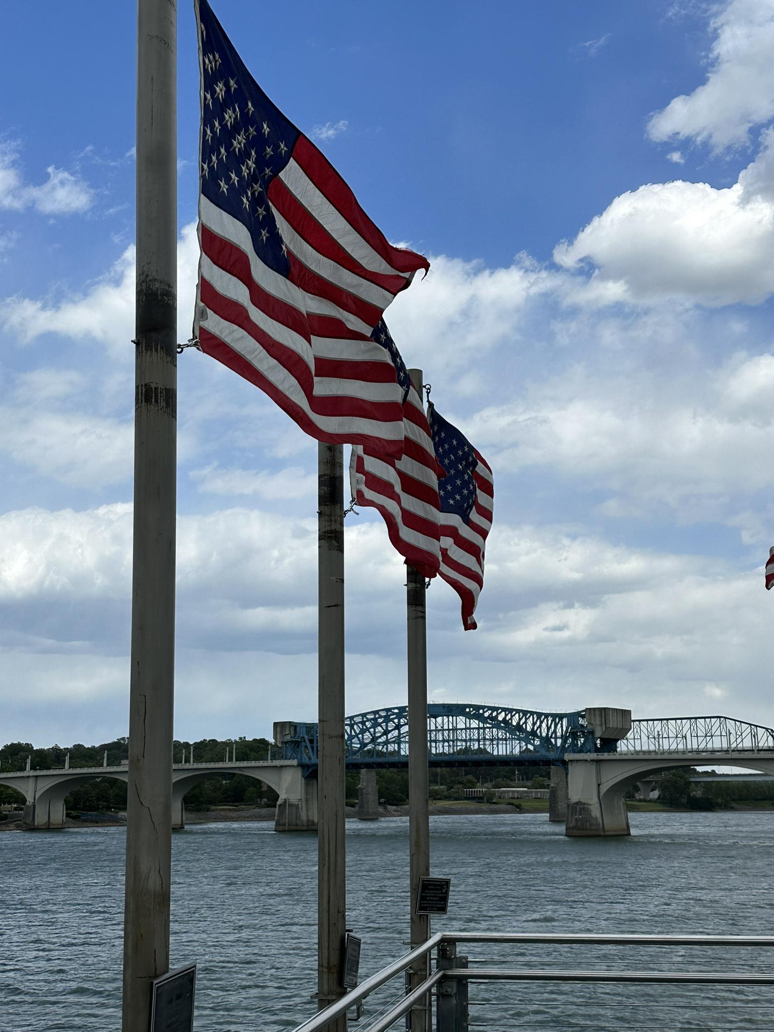 Two American flags wave by a large bridge over water under a cloudy sky.