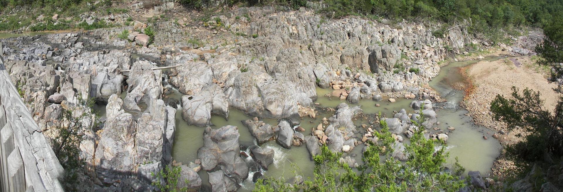 Overhead view of a rocky riverbed with shallow, green-tinged water and sparse vegetation.