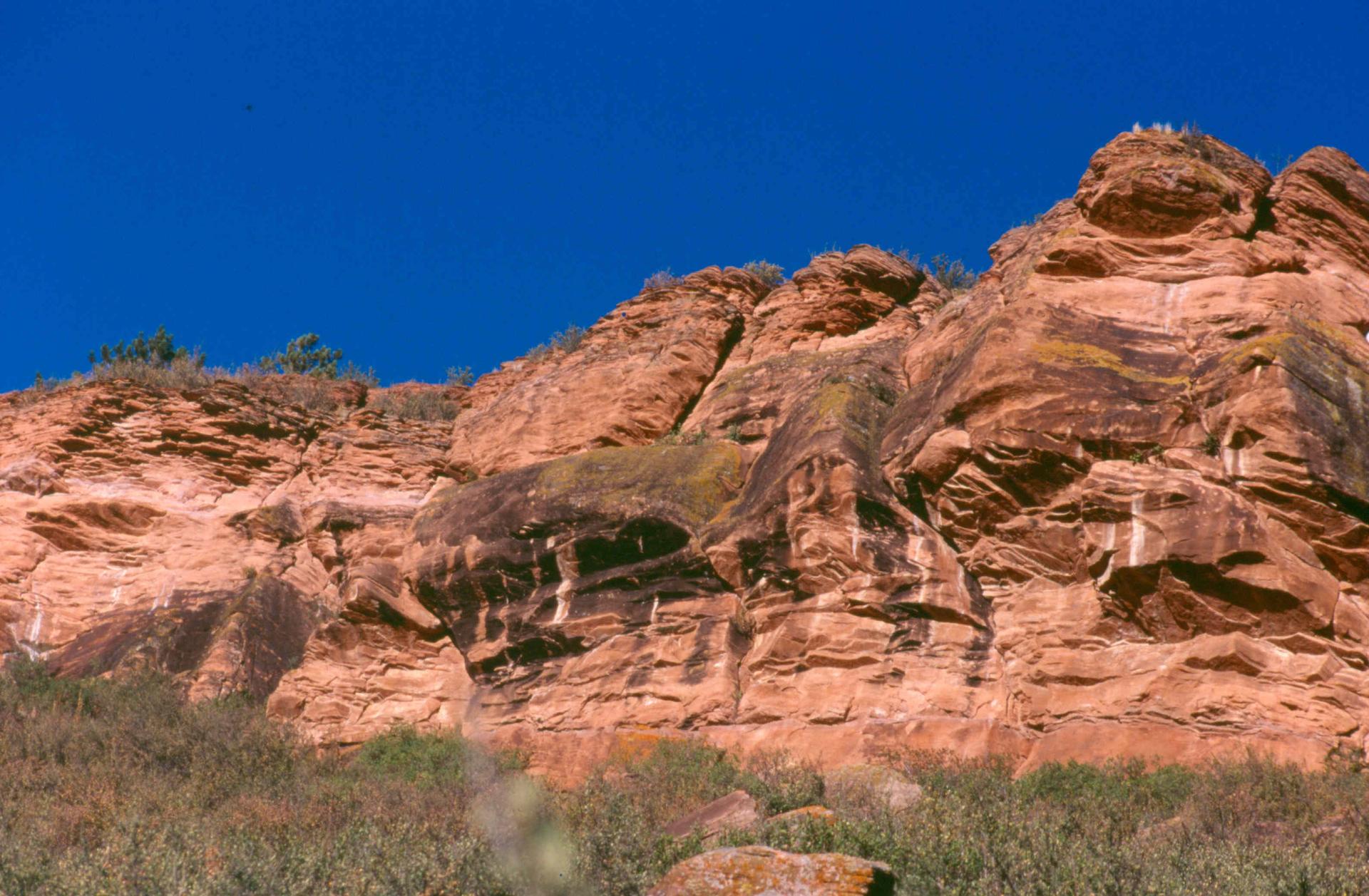 Towering red rock cliff under a bright blue sky with sparse vegetation.
