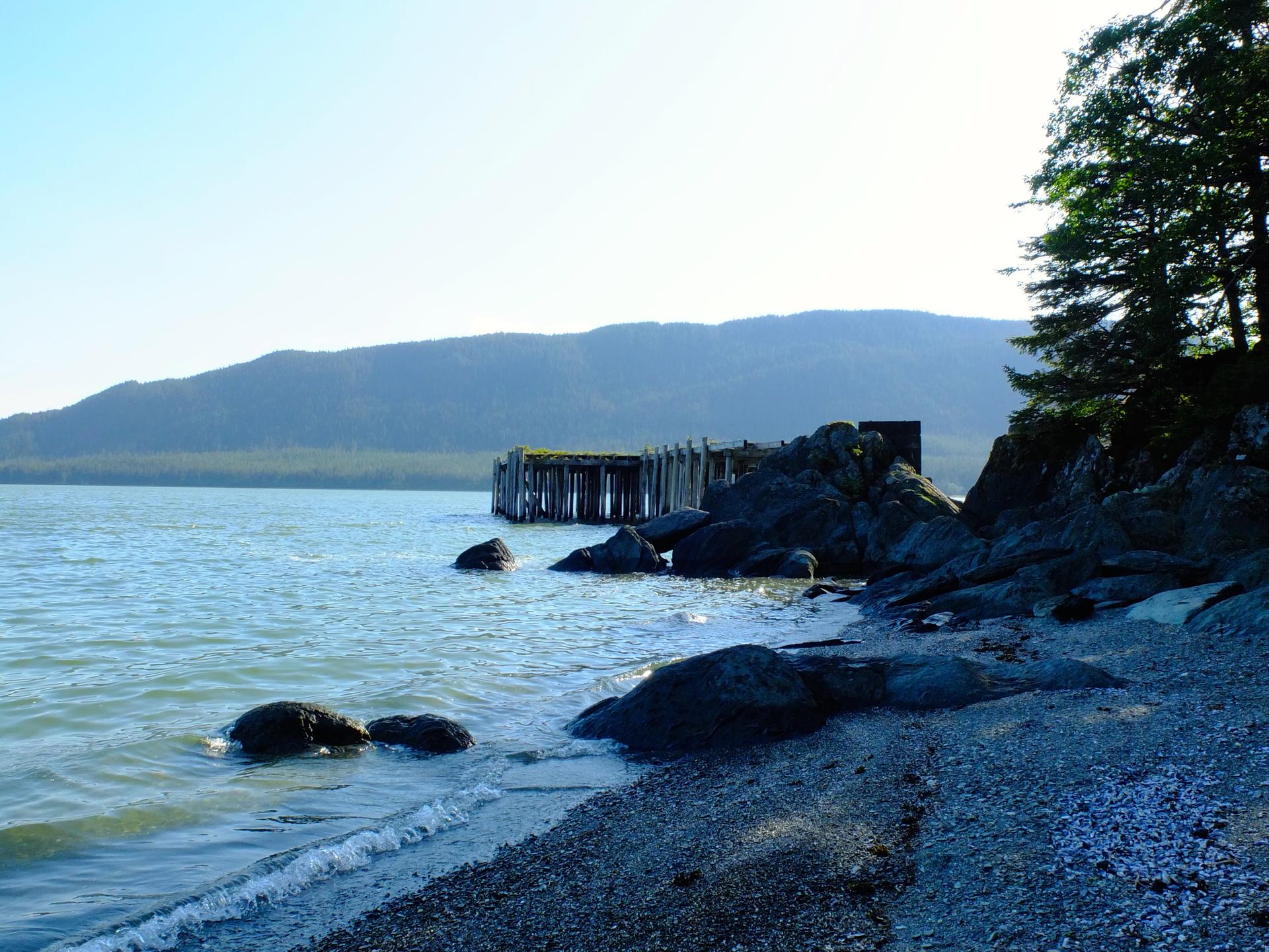 Rocky beach, calm water, old pier, and distant mountains under bright sky.