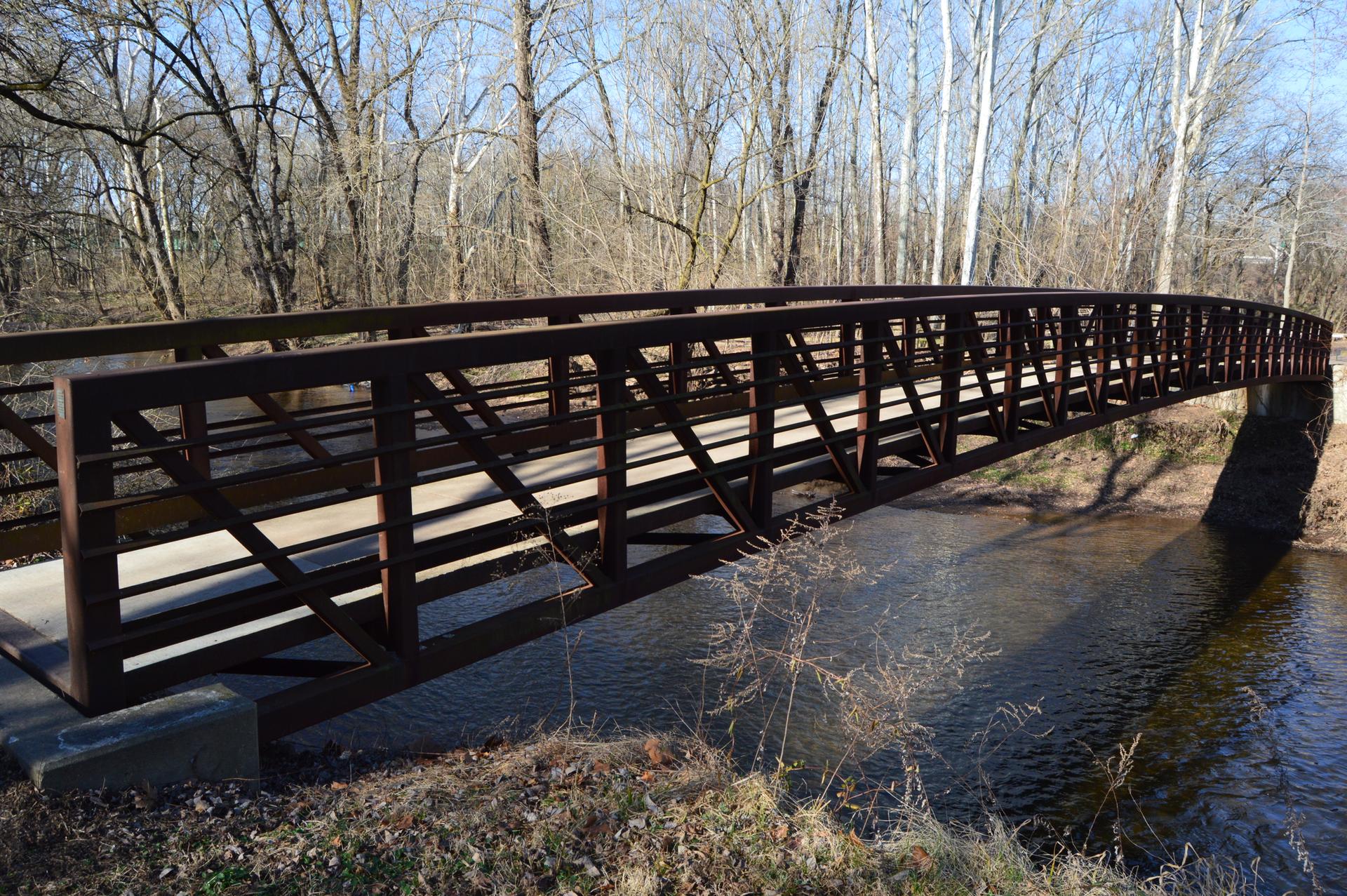 Dark metal truss bridge over a river, surrounded by bare trees in daylight.