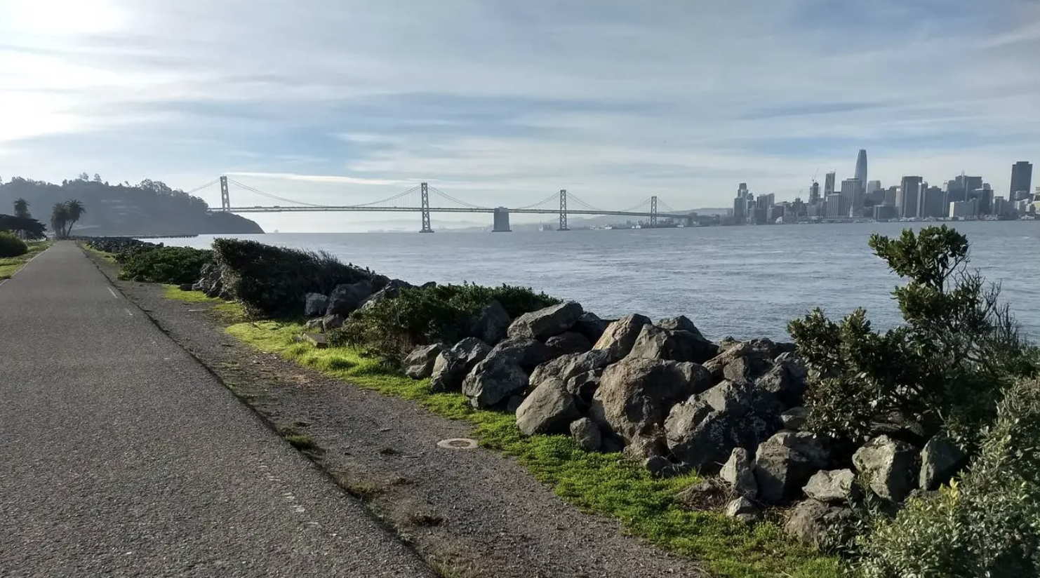 Paved path along a rocky shore with a distant city skyline and bridge.