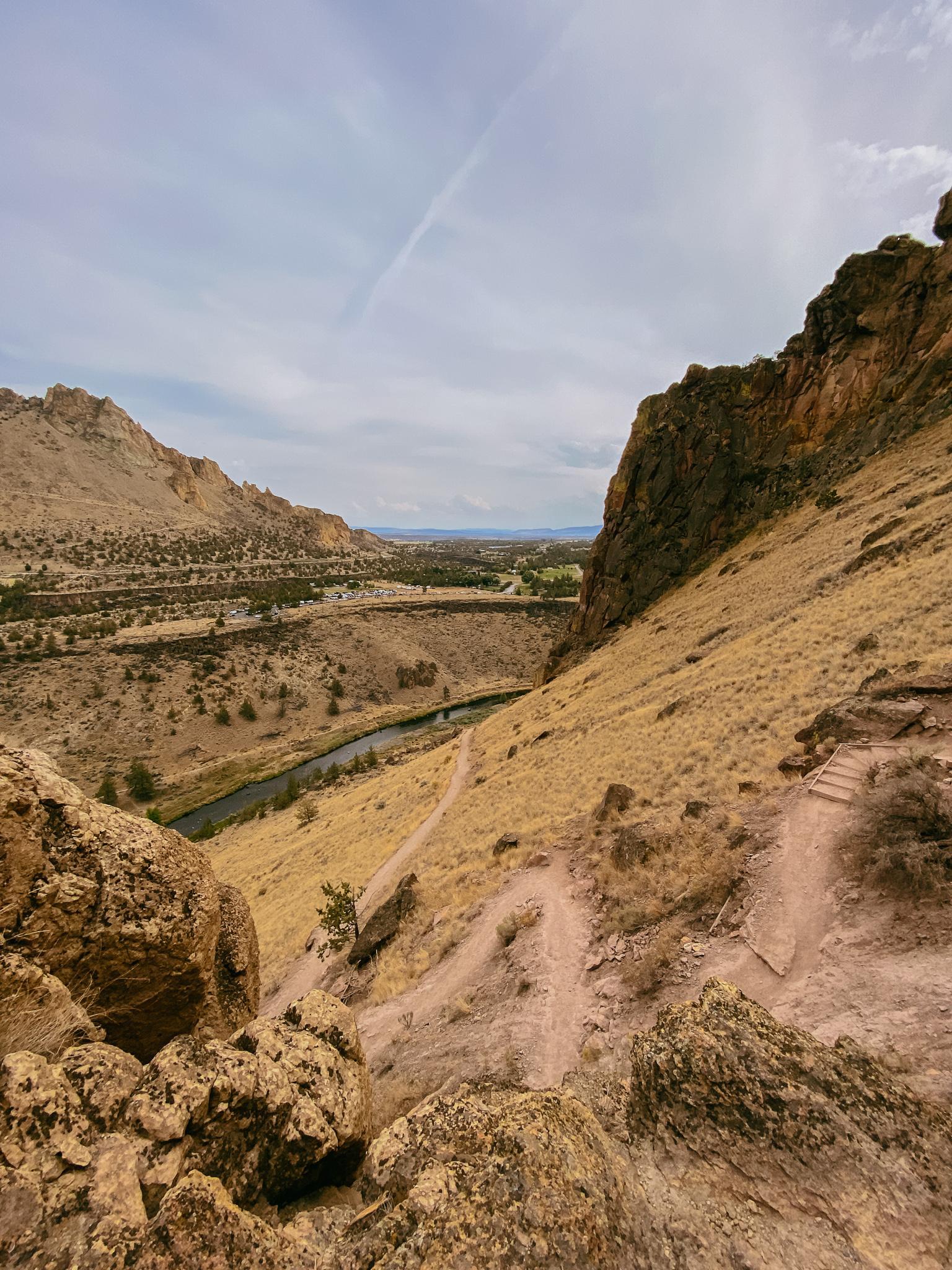 A winding trail descends a steep, dry hillside towards a river below.