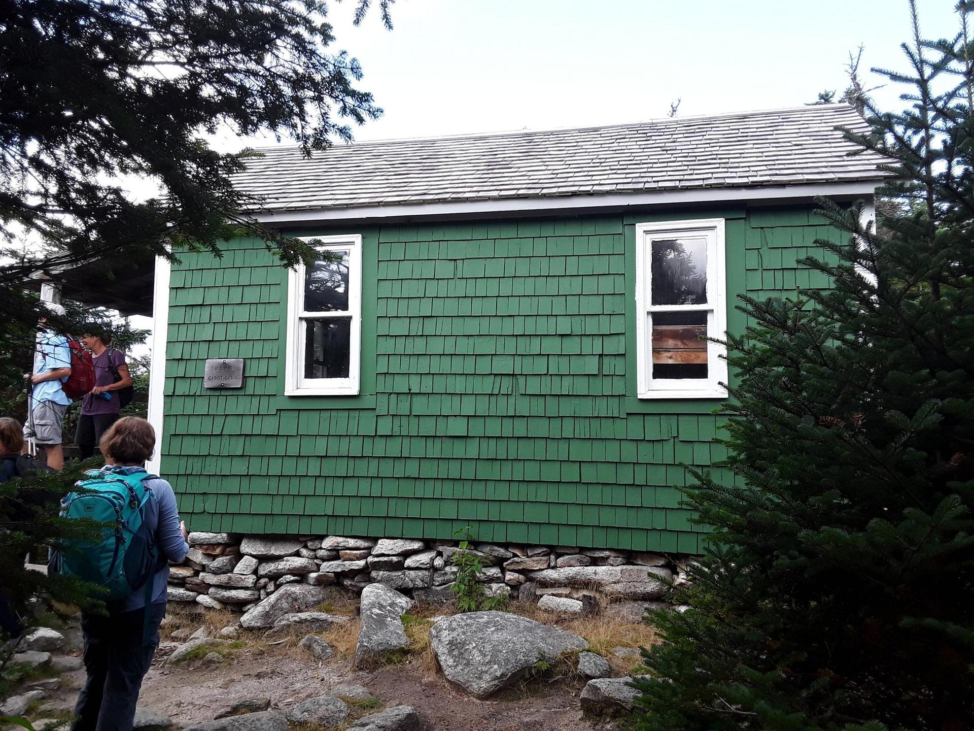People stand near a green shingled cabin with white windows.