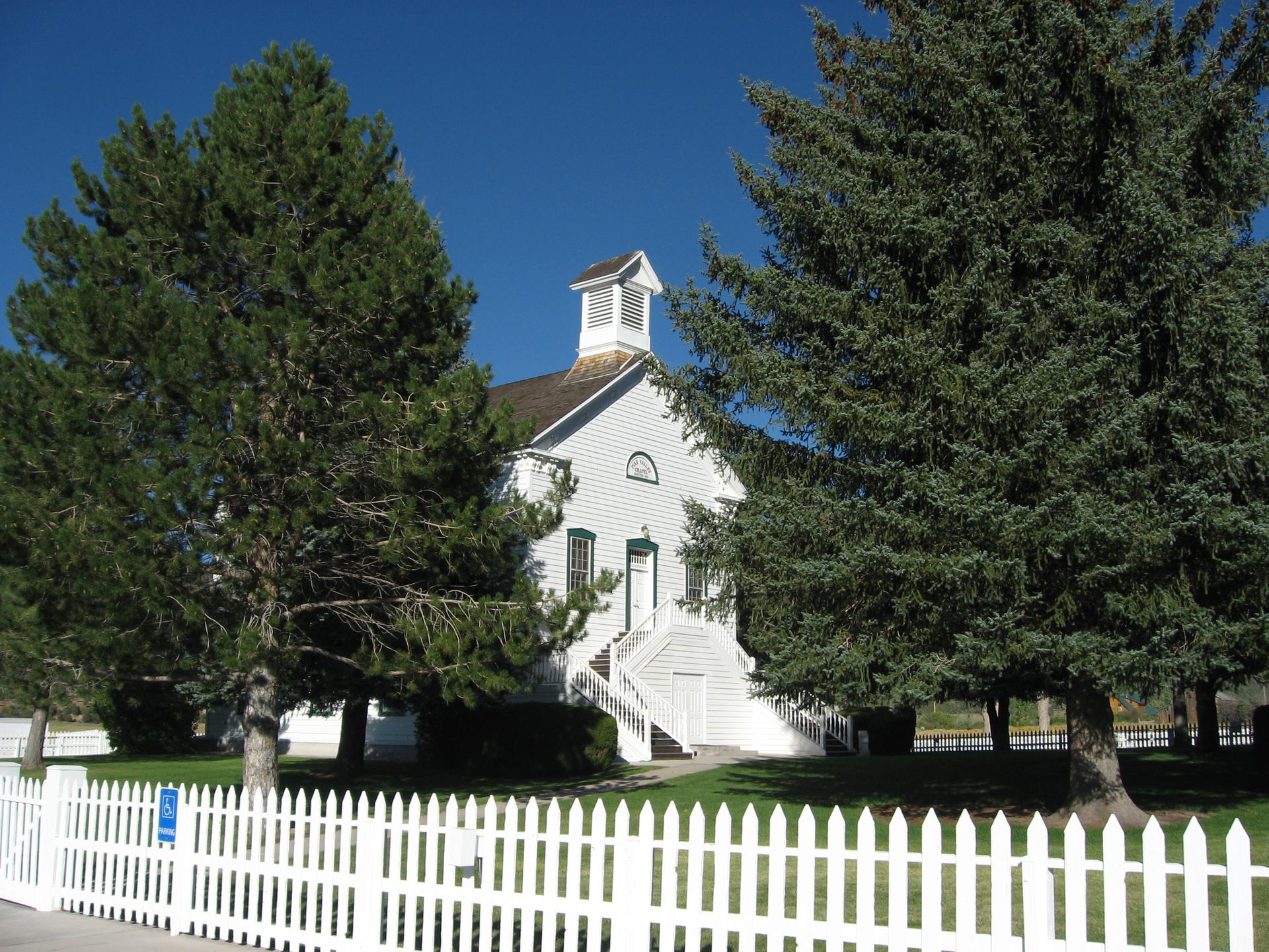 White building with steeple, large evergreen trees, and picket fence on a sunny day.