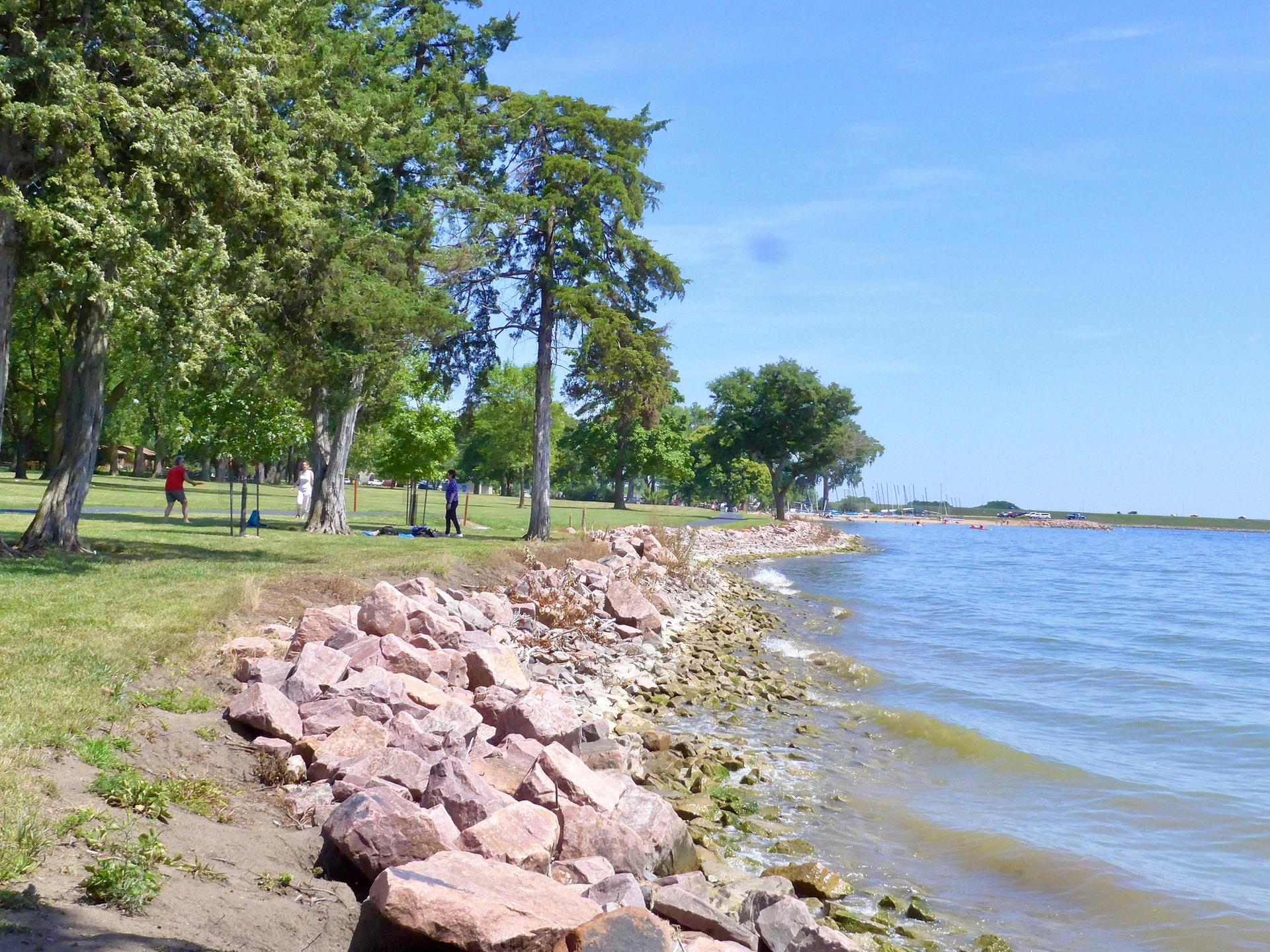 Sunny day with people walking along a rocky lake shore with green trees.