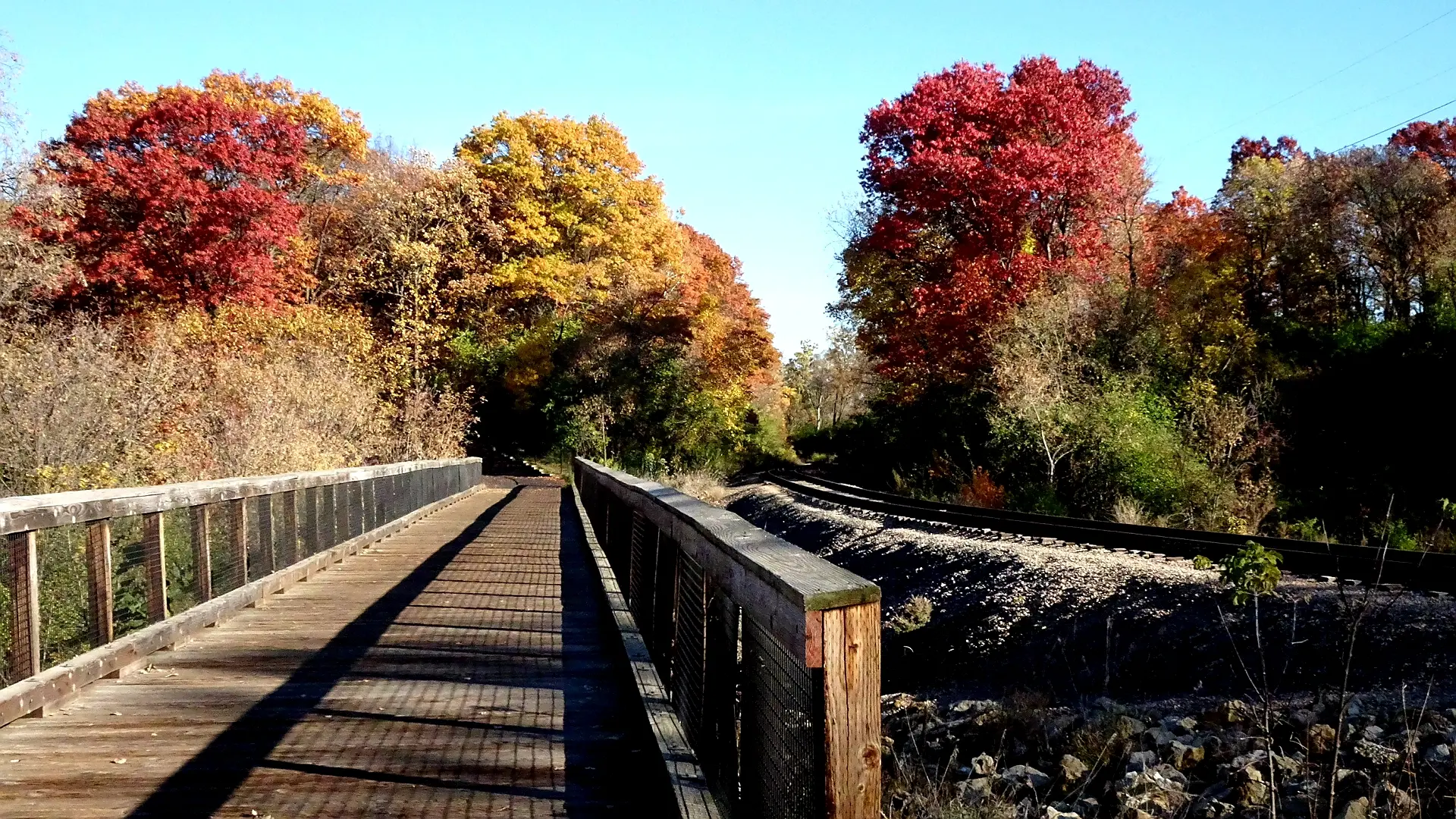 Wooden bridge next to railroad tracks with vibrant autumn trees.