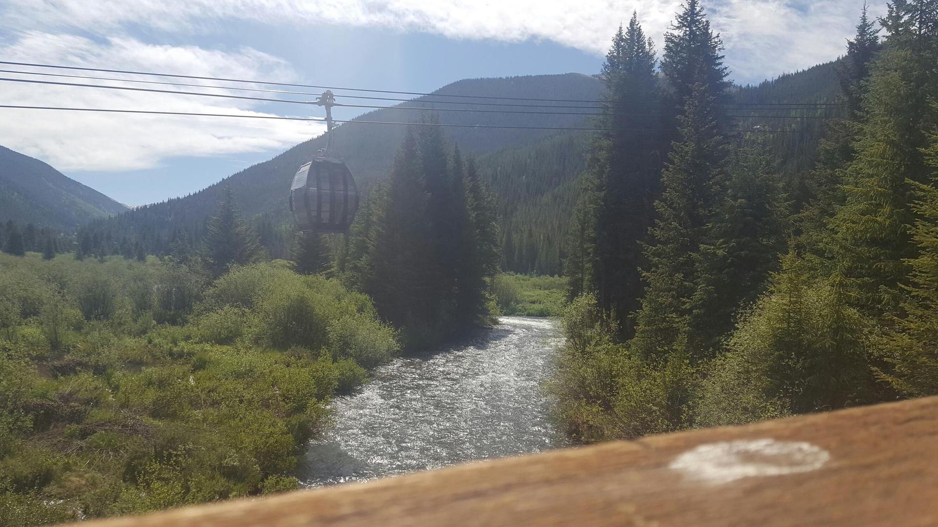 Gondola cabin over a river in a sunny mountain valley with evergreen trees.