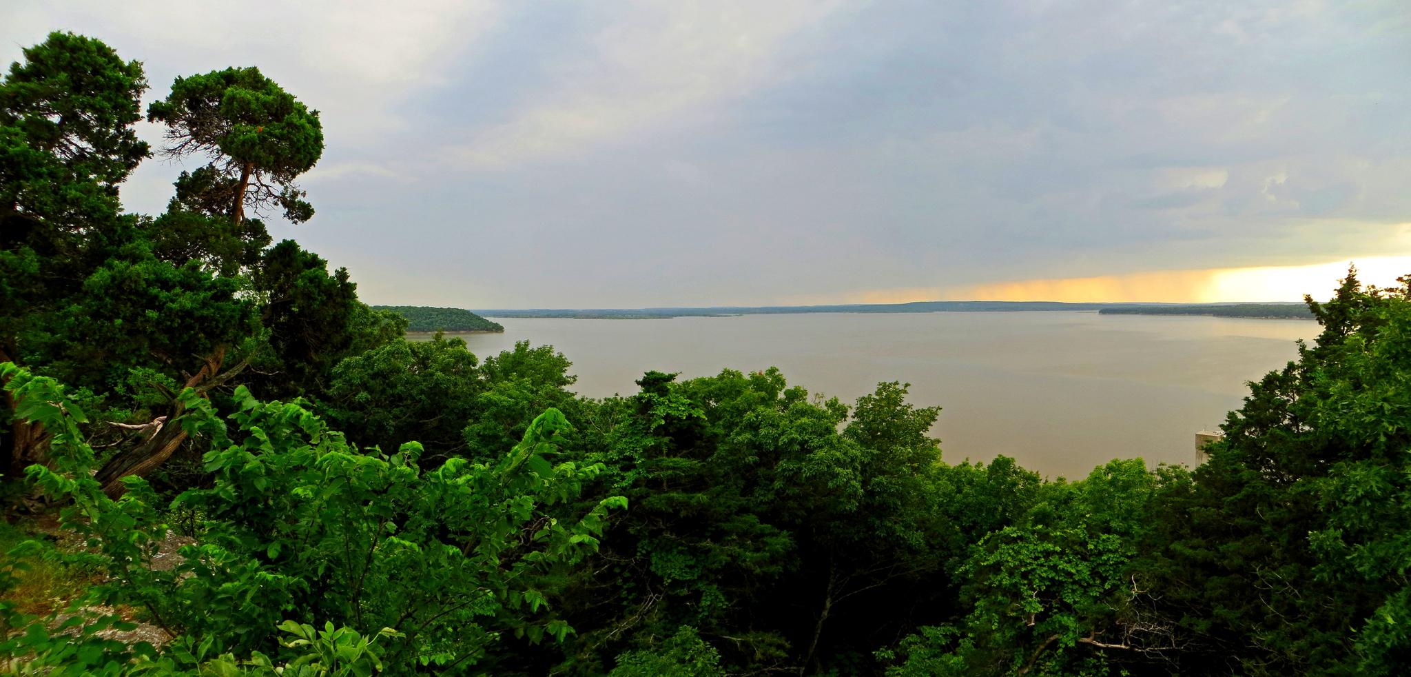 Overlook of a vast lake surrounded by green trees under a cloudy sky.