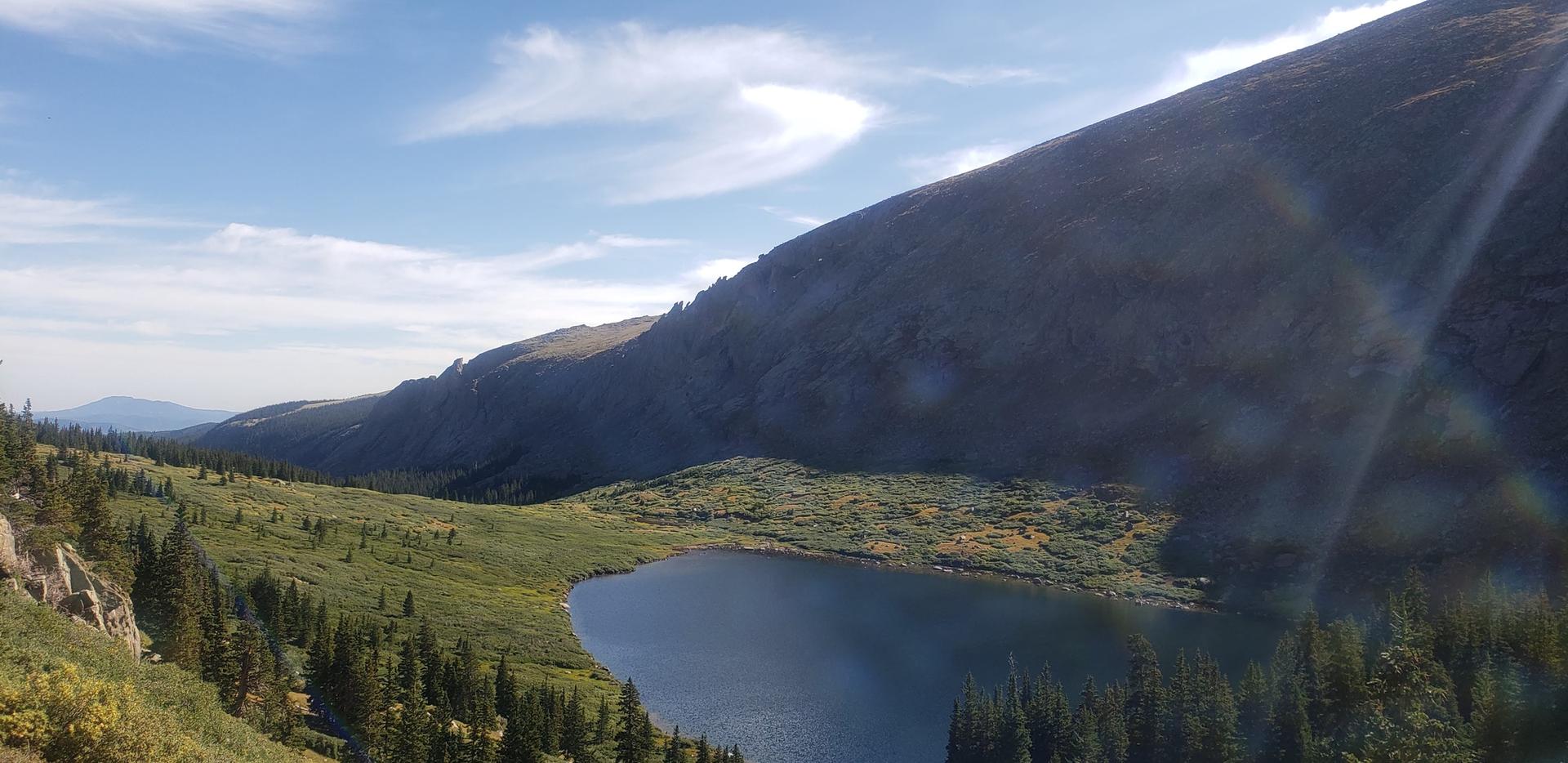 Sunny mountain lake surrounded by green and golden slopes under a blue sky.
