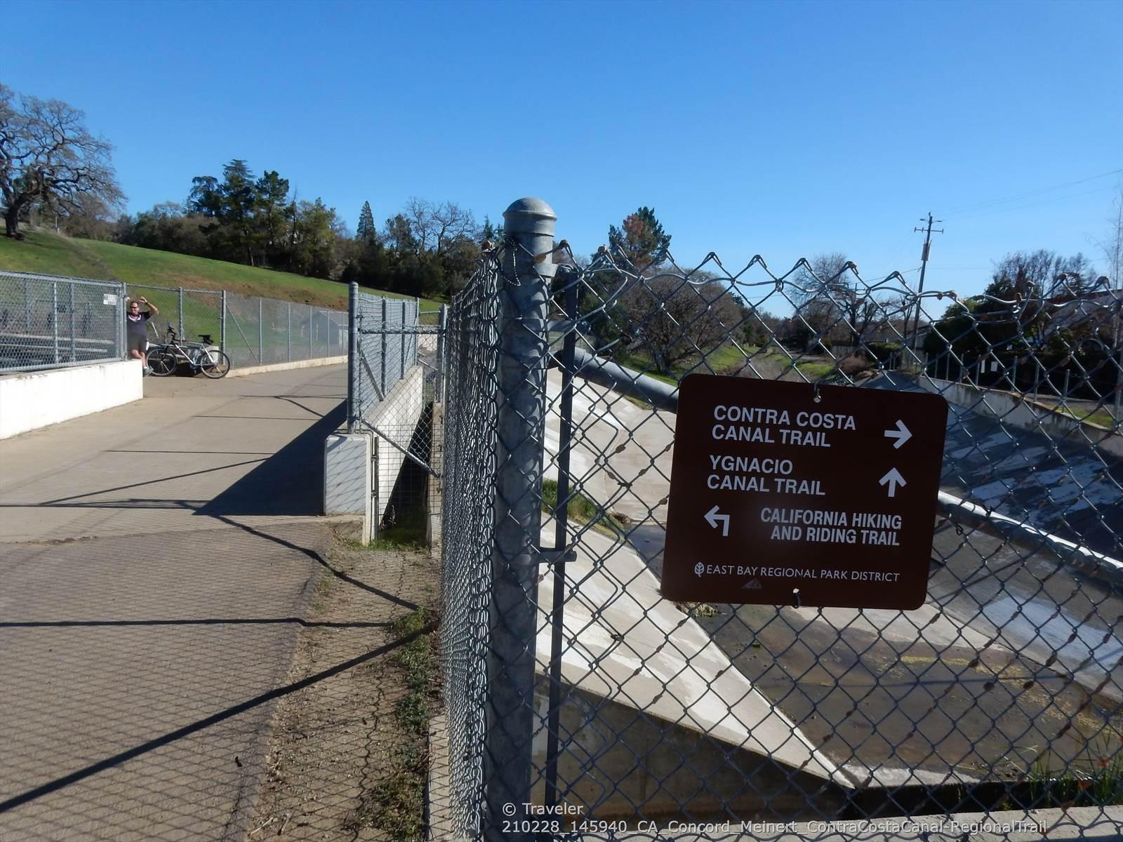 Trail sign on fence by canal, paved path, distant cyclist, bright day.