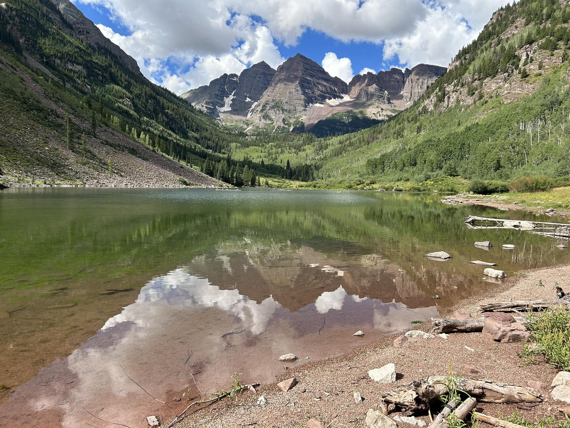 Calm lake reflecting green mountains and clouds under a bright sky.
